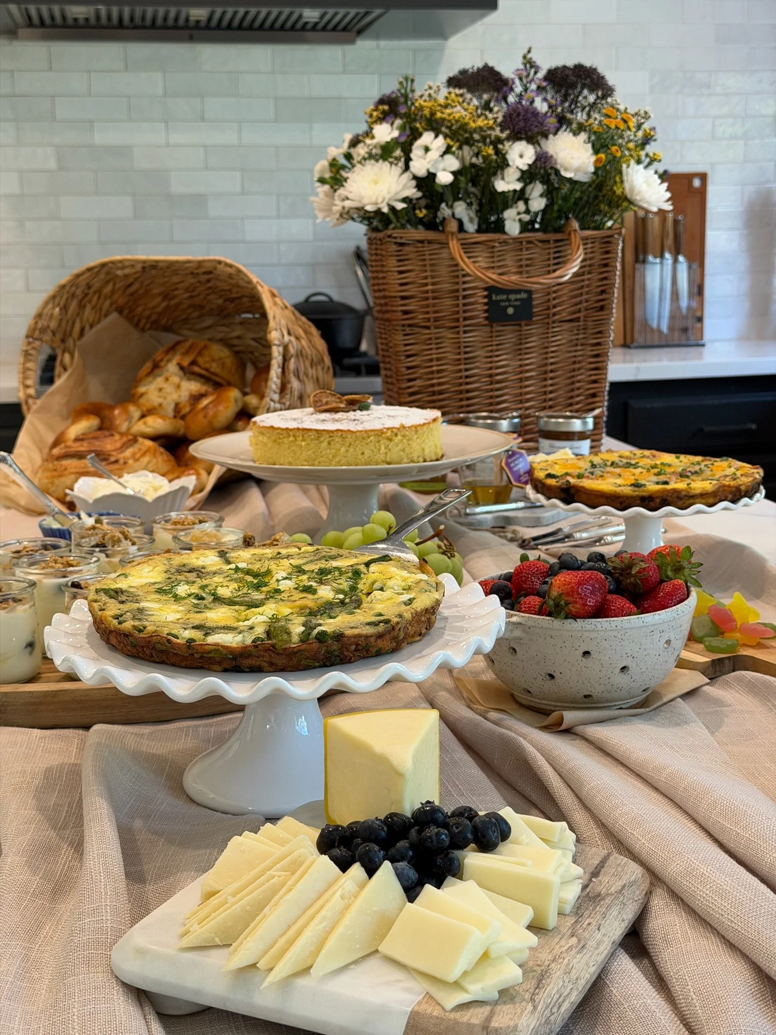 A spread of breakfast foods on a table, including cheeses, grapes, berries, a quiche, a cake, pastries, and a bowl of fruit, with a large bouquet of flowers in the background.