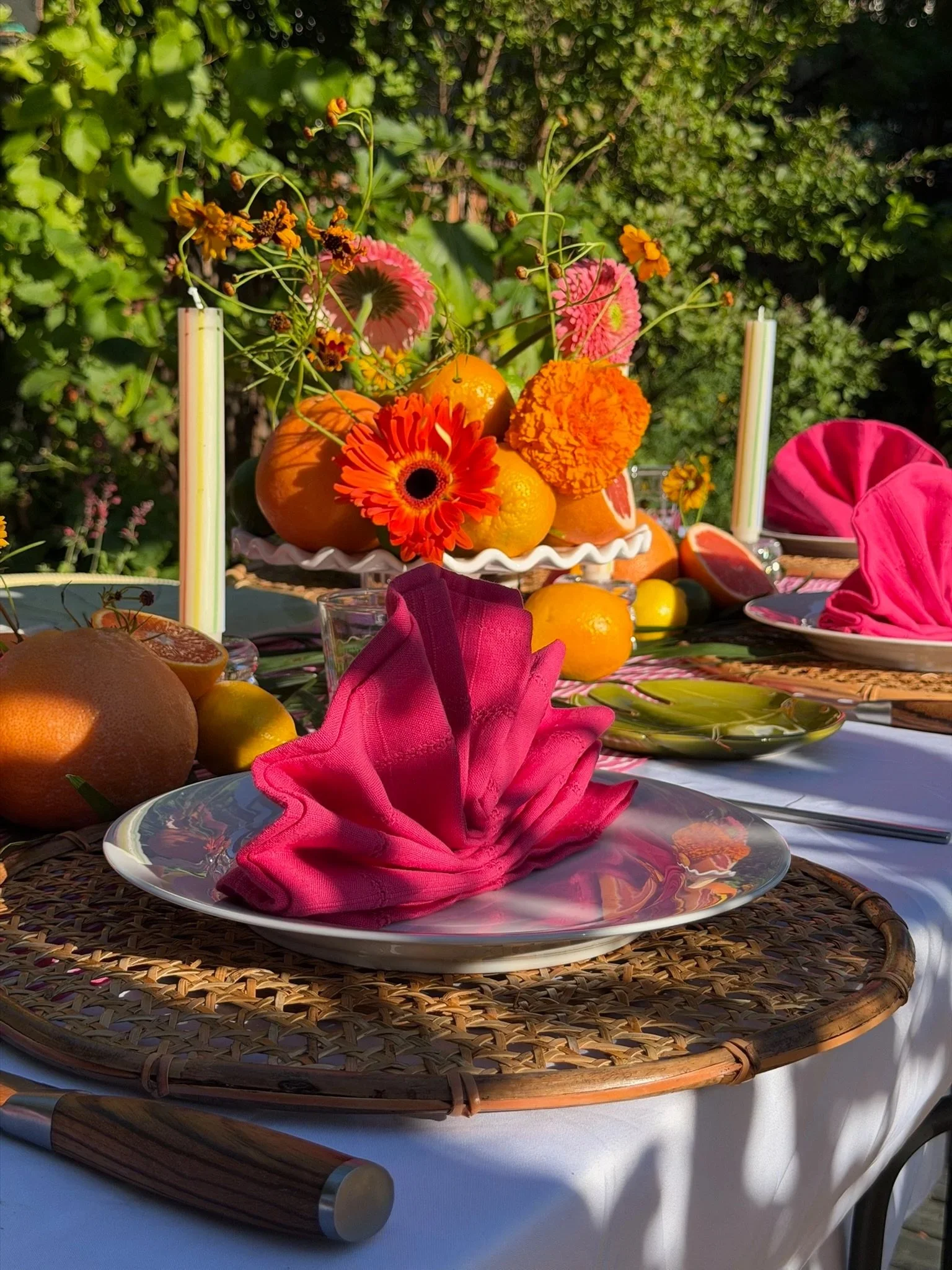 A table set outdoors with a wicker placemat, pink napkin, and white plate holding a pink cloth. On the table, there is a fruit centerpiece with oranges, grapefruits, and pink daisies. Pink candles and additional plates with fruit are also visible, with lush green foliage in the background.