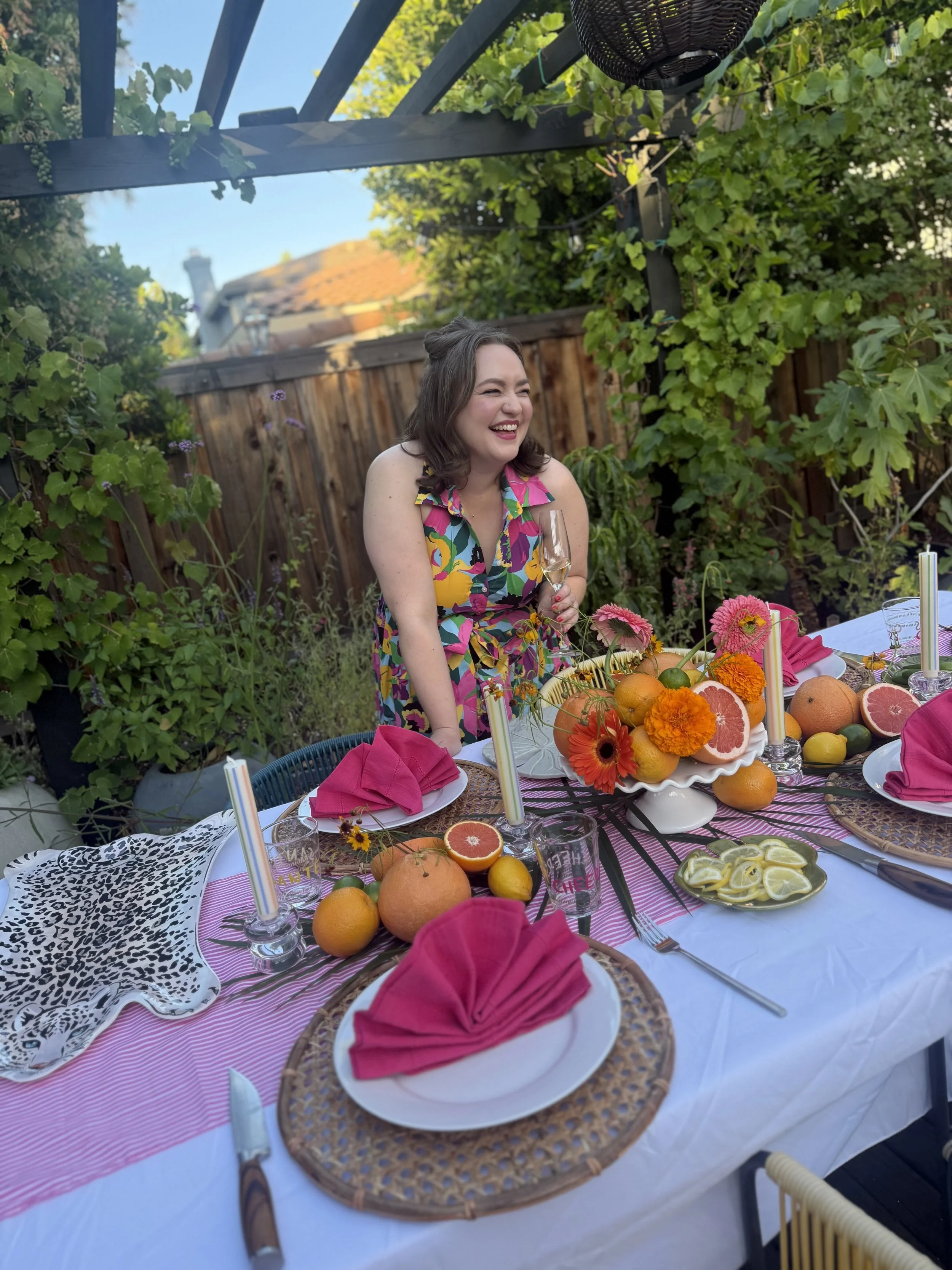 A woman in a colorful floral dress sitting at an outdoor table, smiling and holding a glass of champagne, with a fruit centerpiece including grapefruit, oranges, lemons, and marigolds, set for a celebration or gathering in a garden with greenery and a wooden fence.