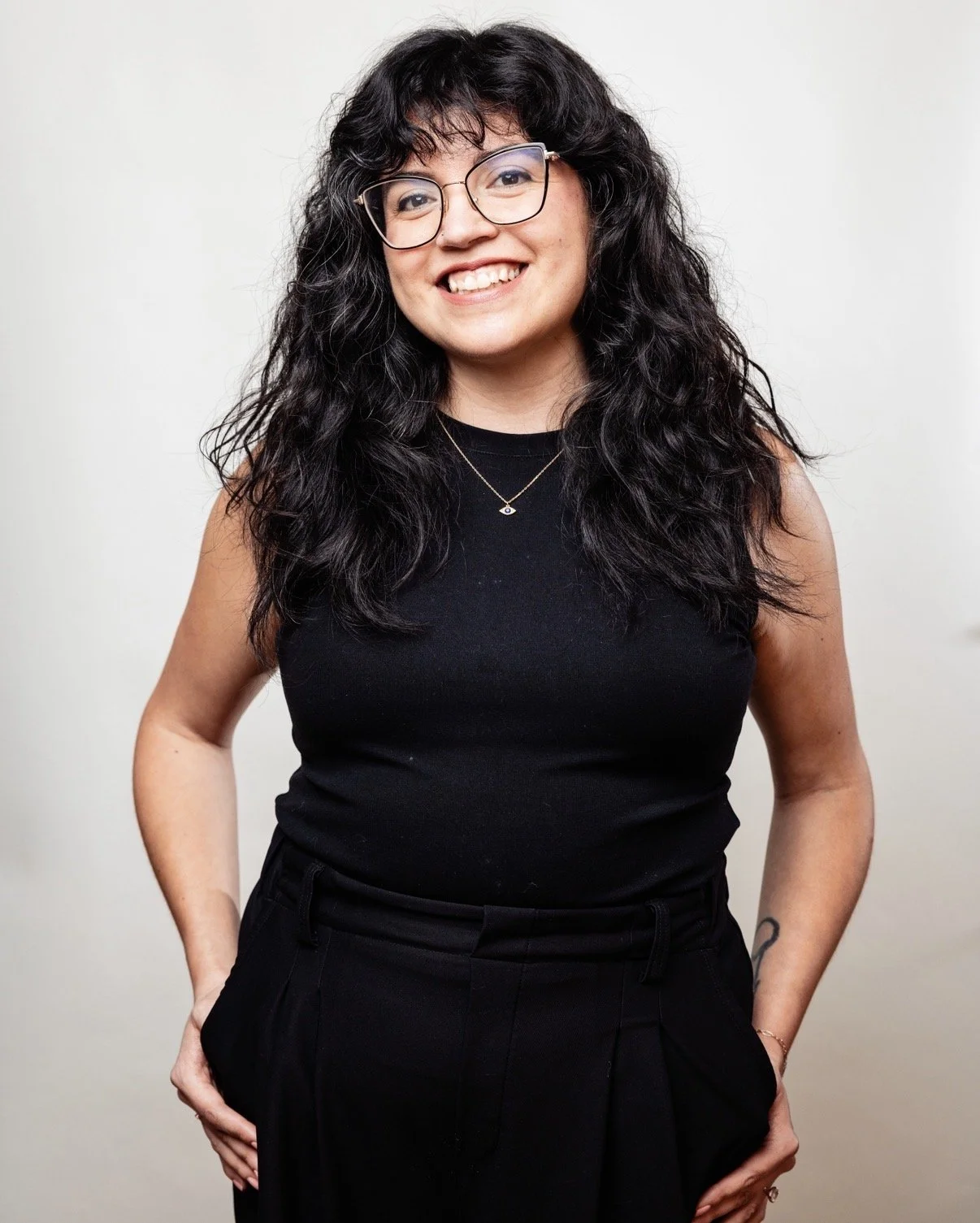 A woman with curly black hair and glasses smiling, wearing a black sleeveless top and black pants against a plain white background.
