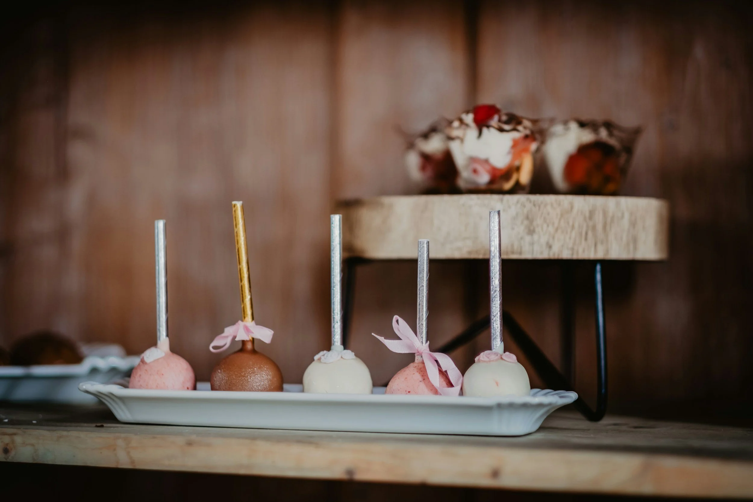 Colorful cake pops with pink, white, and brown coatings, some decorated with bows, on a white tray in front of a wooden background, with a blurred layered dessert in a bowl above.