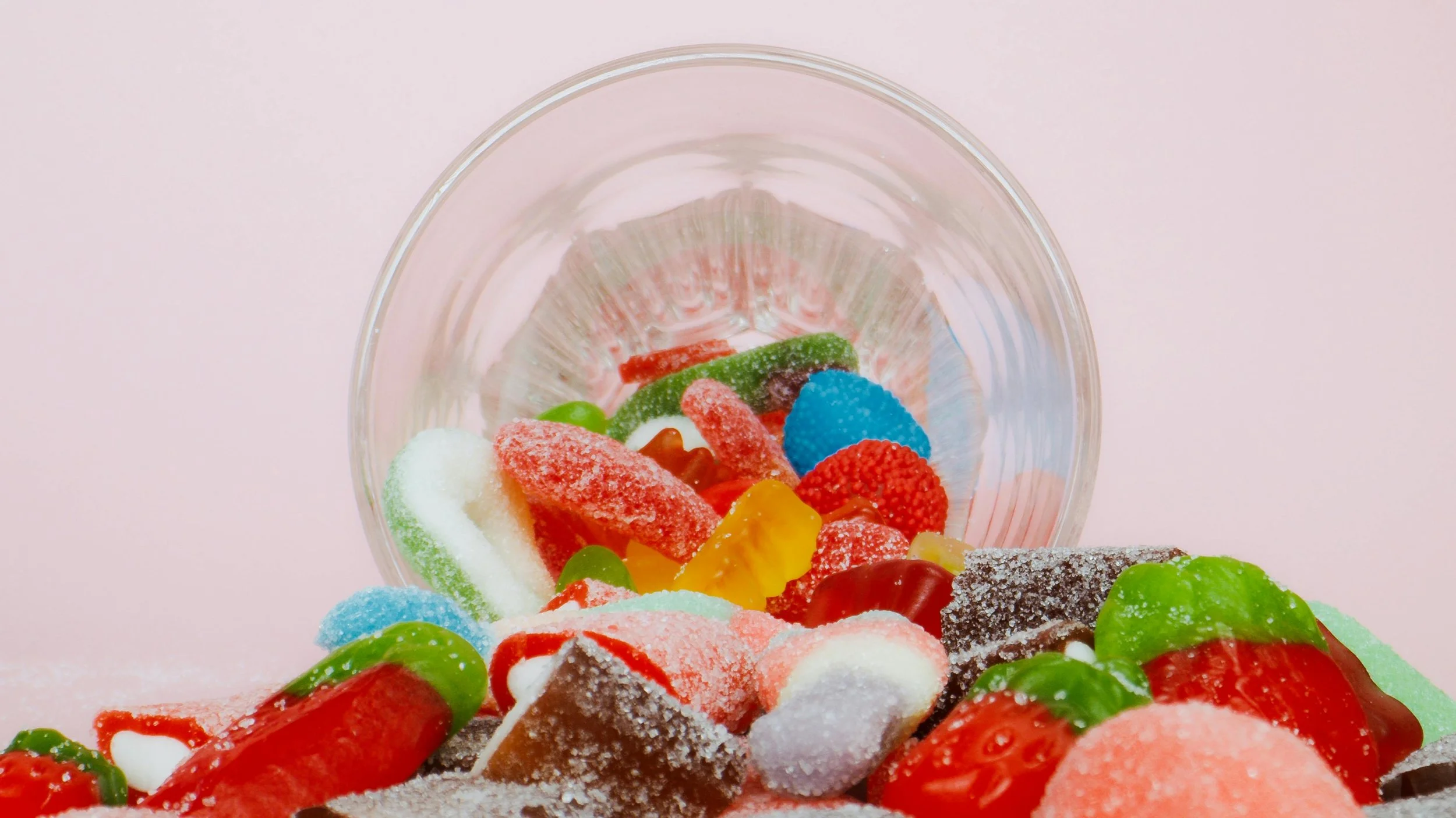 Assorted colorful candies spill out of a glass container onto a pastel pink surface.