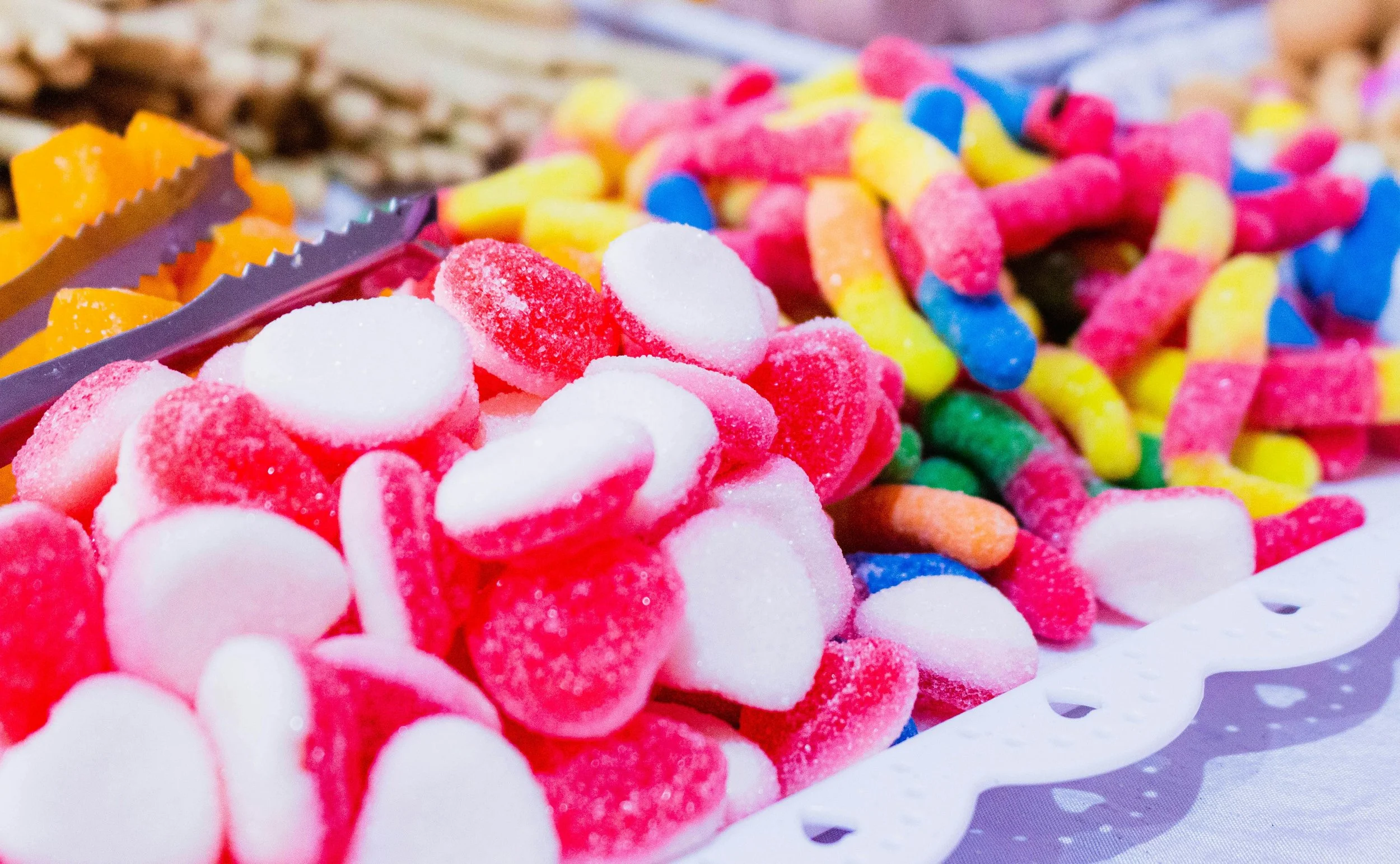 Colorful gummy candies, including red and white heart-shaped gummies, rainbow worms, and yellow-orange fruit-shaped gummies on a white tray.