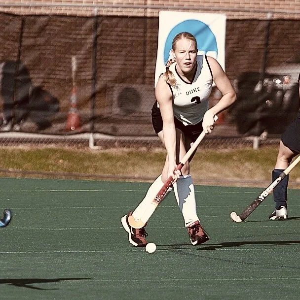 Gracie Sorbello Cole playing field hockey on an outdoor turf field at Duke University, years before playing on the USA masters field hockey national team, holding a field hockey stick, and dribbling a ball during a game.