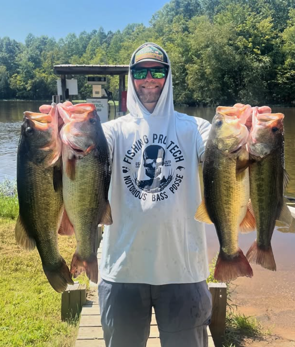 Man holding two large bass fish in each hand outdoors near the water on a sunny day.