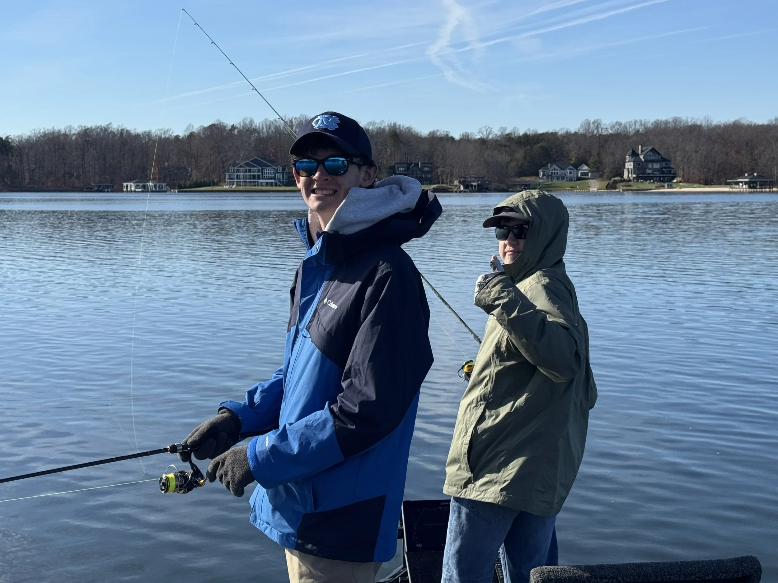 Two males fishing on a boat in a lake with modern houses on the shoreline and a wooded hill in the background, under a clear blue sky.