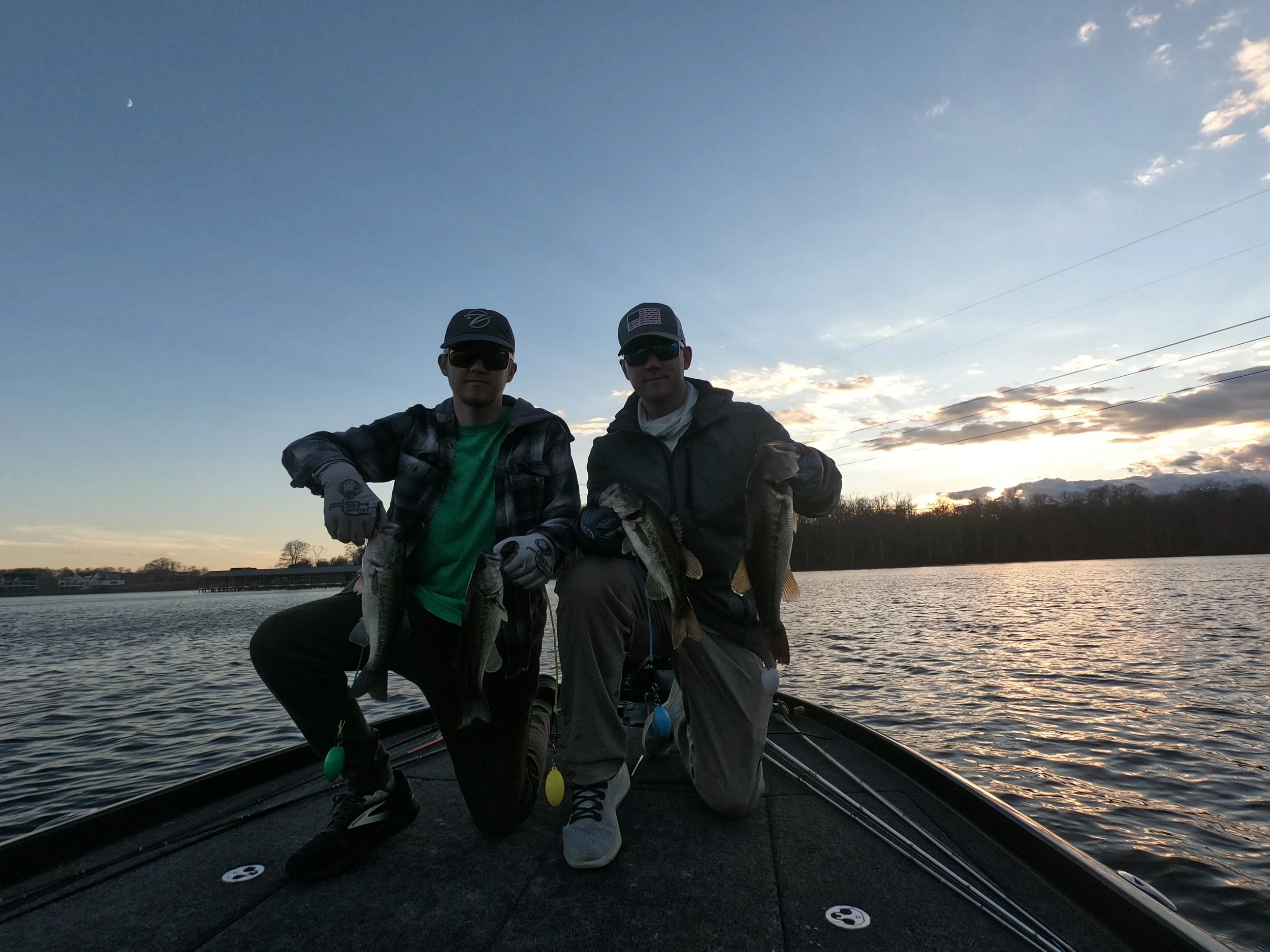 Two men on a boat holding fish they caught, with water and sky in the background at sunset.