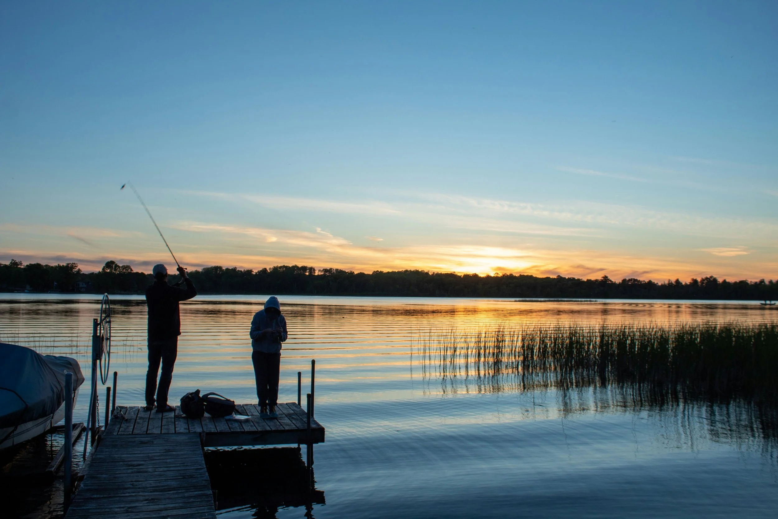 Two people fishing off a dock at sunset on a calm lake, with trees in the background and a partly cloudy sky.