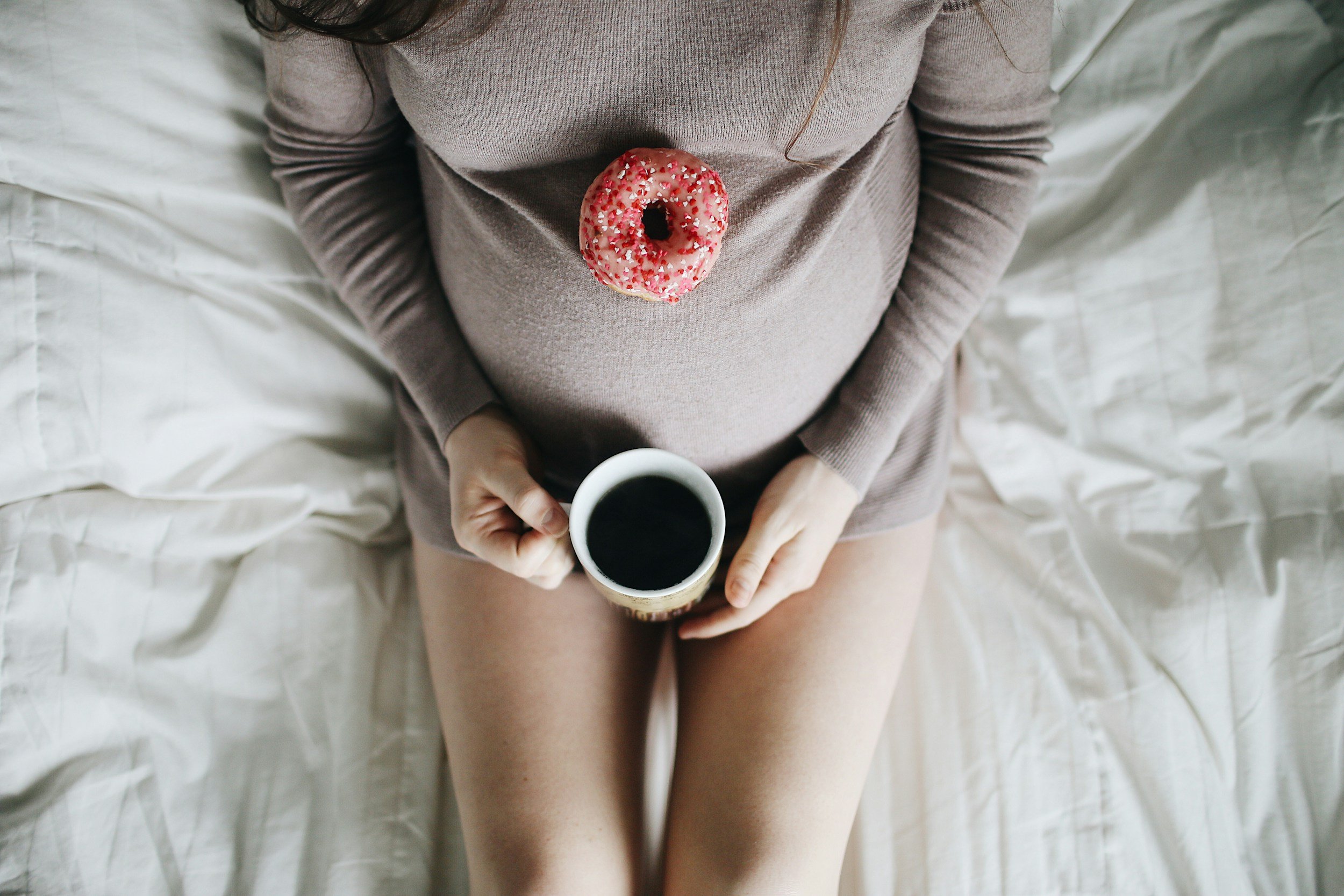 Une femme enceinte assise sur un lit, tenant une tasse de café, avec un beignet rose recouvert de paillettes posé sur son ventre.