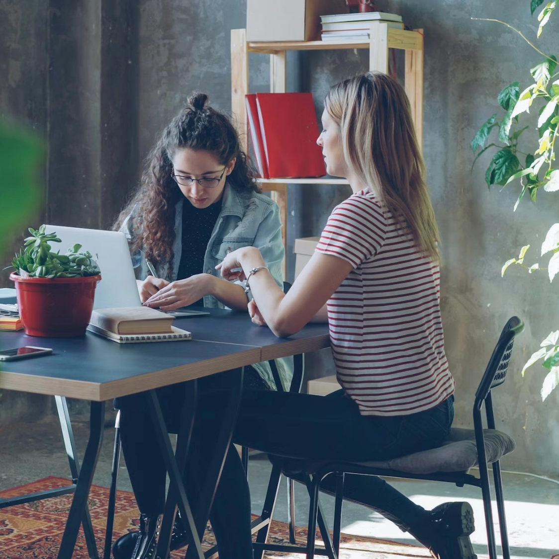Deux femmes assises à une table de travail en intérieur, discutant et travaillant, avec des livres, un ordinateur portable, une plante en pot, et une étagère en bois en arrière-plan.