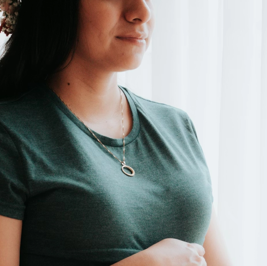 Une femme porte un t-shirt vert foncé et une chaîne avec un pendentif ovale, près d'une fenêtre avec des rideaux blancs.