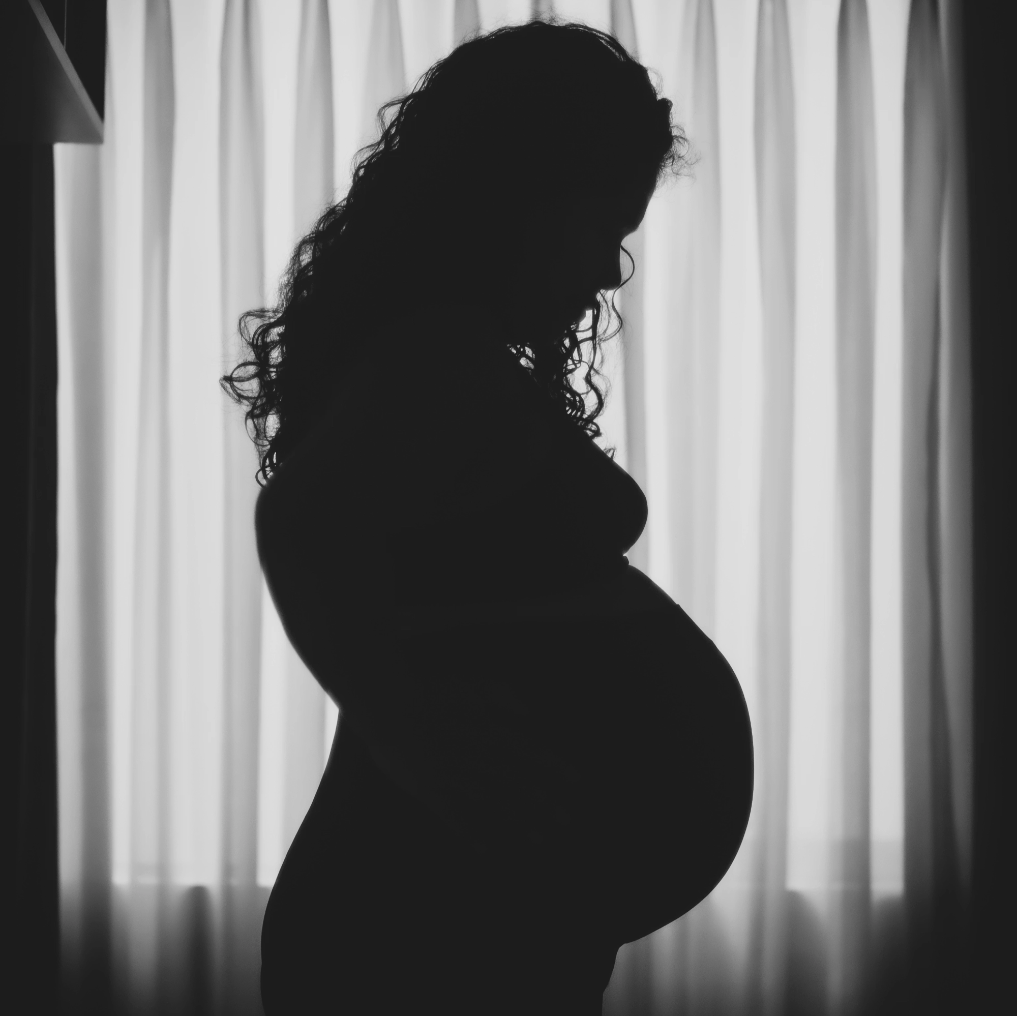 Silhouette d'une femme enceinte regardant vers le bas, avec des rideaux en arrière-plan.