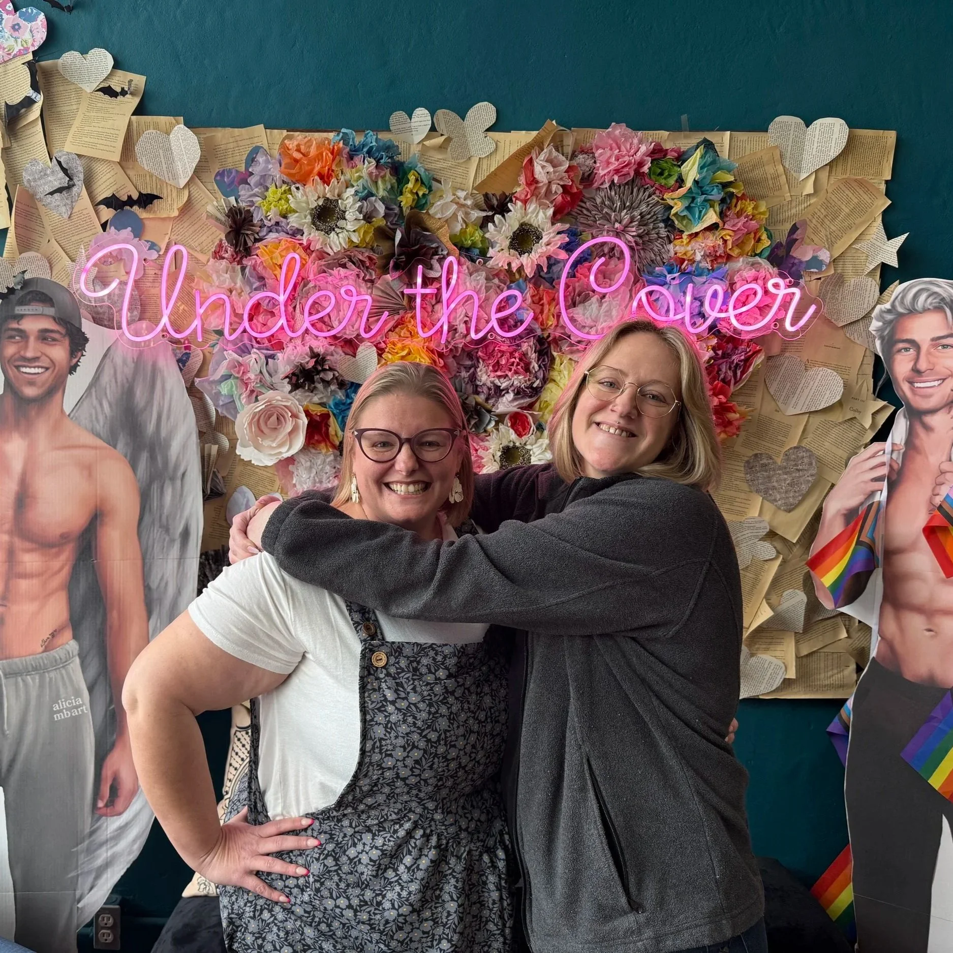 Two women hugging in front of a colorful backdrop with paper flowers, books, and cutouts of shirtless men, including one with rainbow pride accessories, with a neon sign that says "Under the Cover."