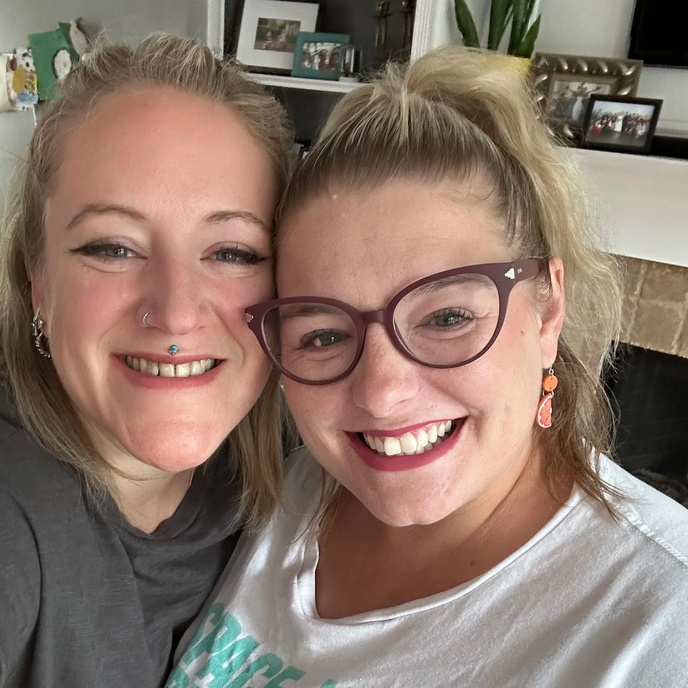 Two women smiling and taking a close-up selfie indoors, with a background of a shelf and framed pictures.