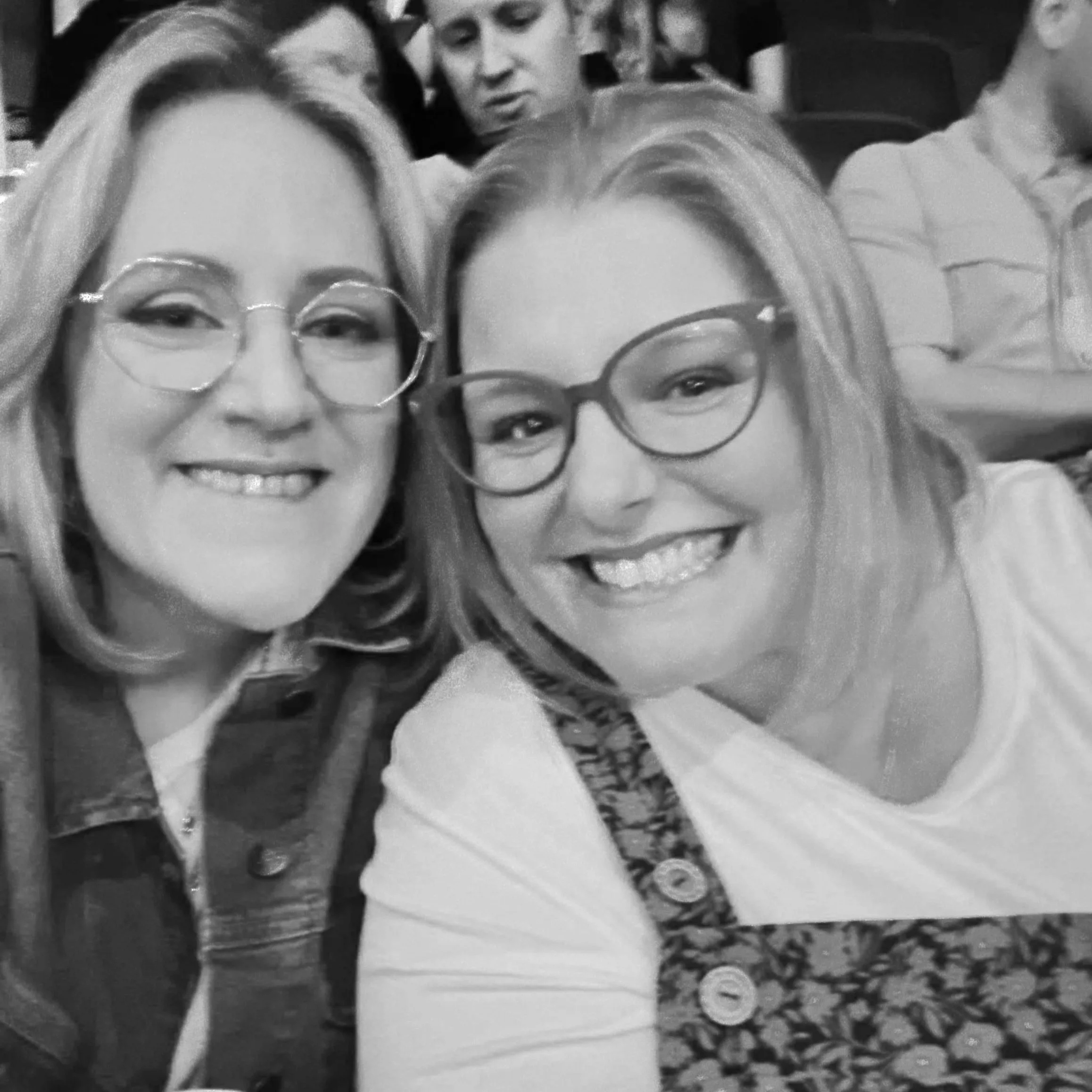 Two women with glasses smiling at the camera, sitting in a crowded indoor event, with a man visible in the background.