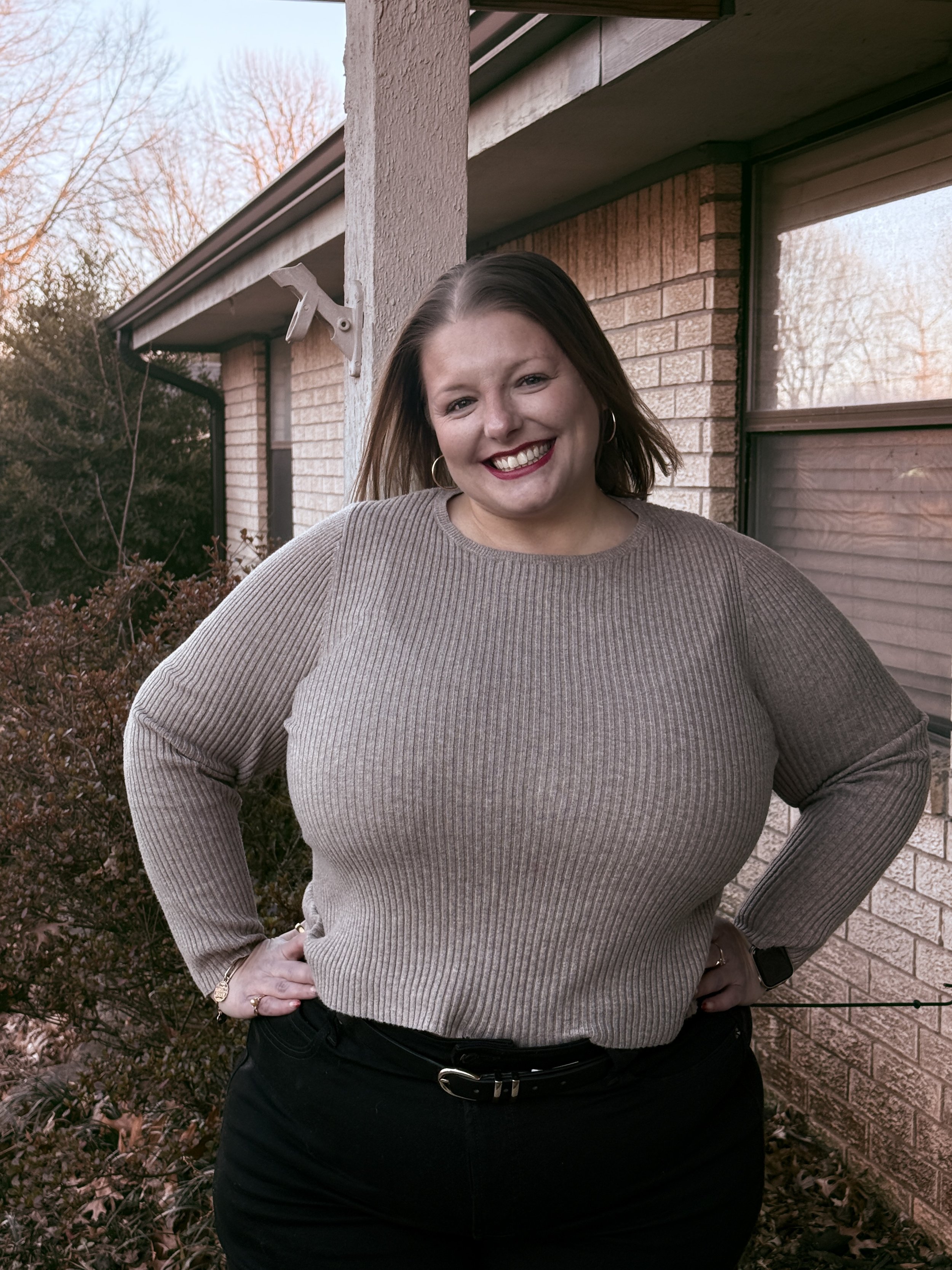 A woman with shoulder-length brown hair smiling, standing outdoors in front of a brick house with a rain gutter, during late afternoon or early evening. She is wearing a beige ribbed sweater and black pants.