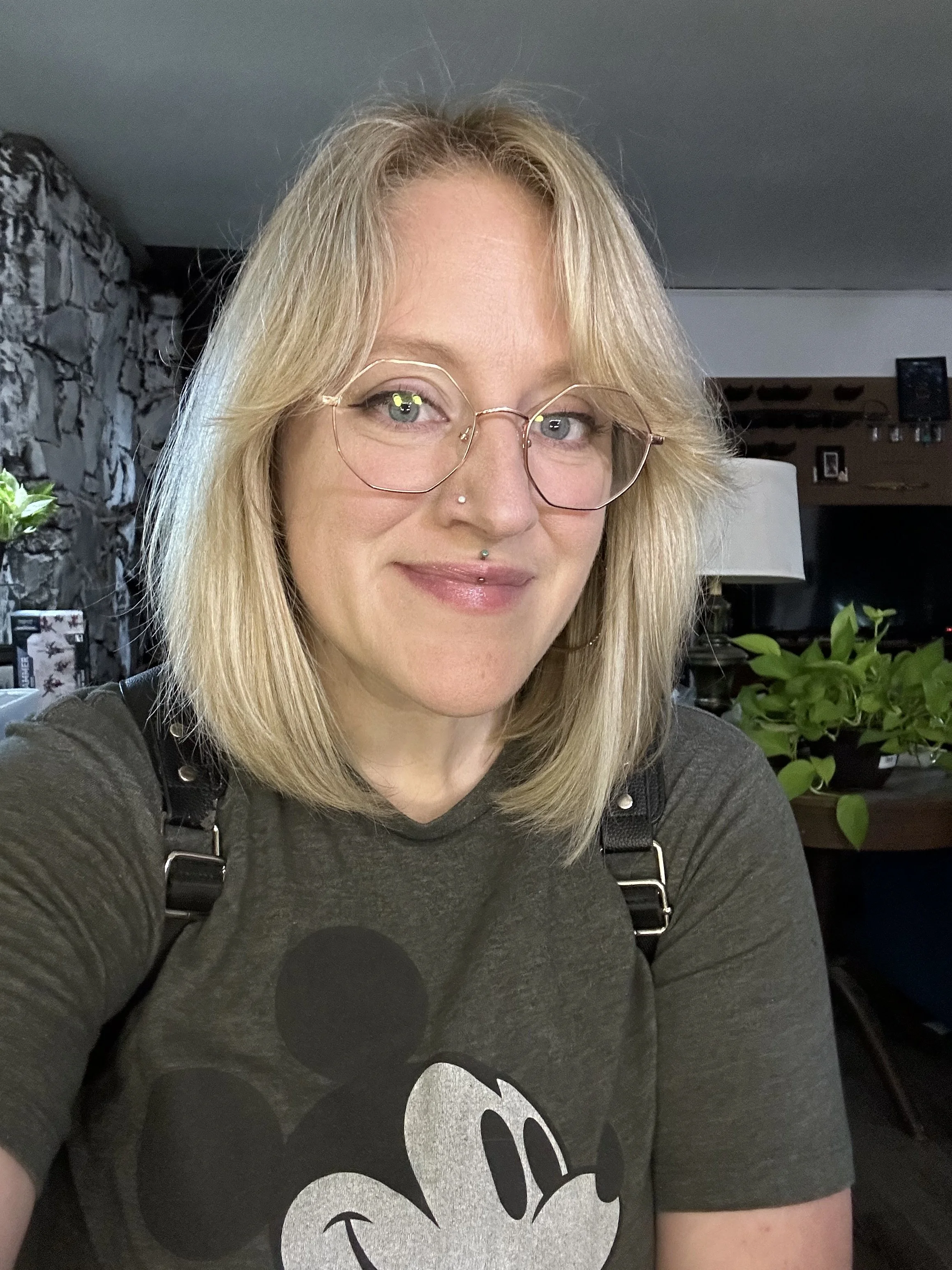 A woman with blonde hair, glasses, and a nose piercing smiling at the camera inside a home with stone walls, a lamp, potted plant, and framed pictures in the background.