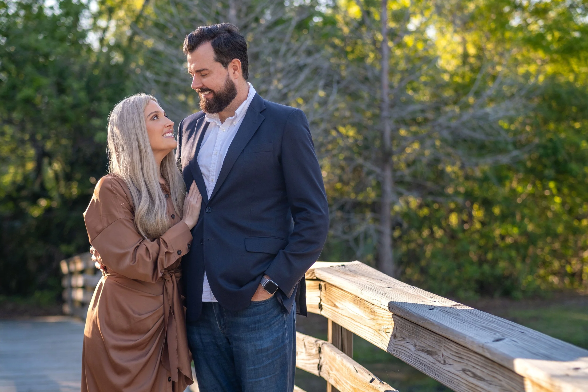 A man and woman stand close together on a wooden bridge, smiling at each other, with a background of trees and sunlight.