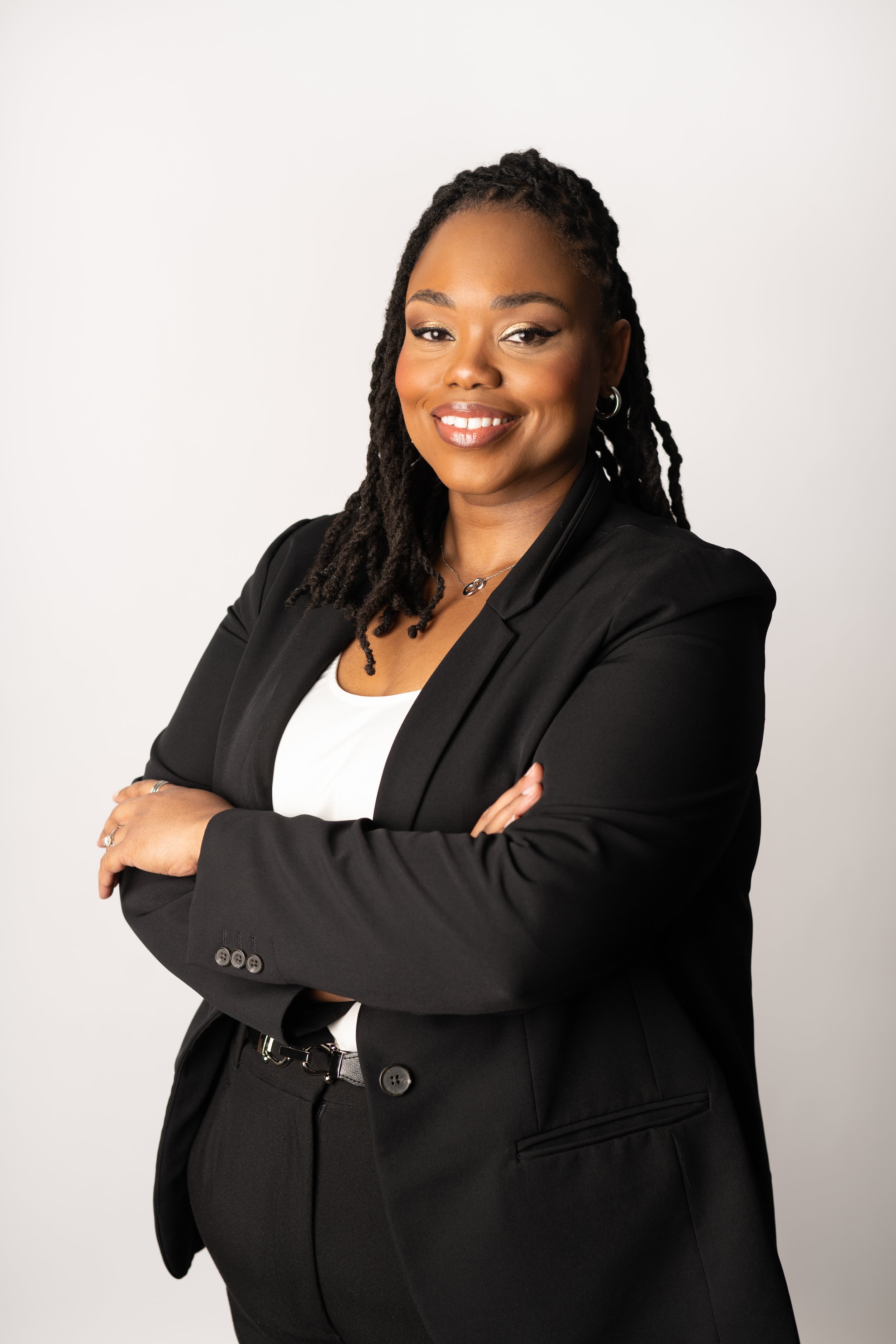 Professional Black woman with black hair in locs wearing a black blazer and white top, smiling with crossed arms against a white background.