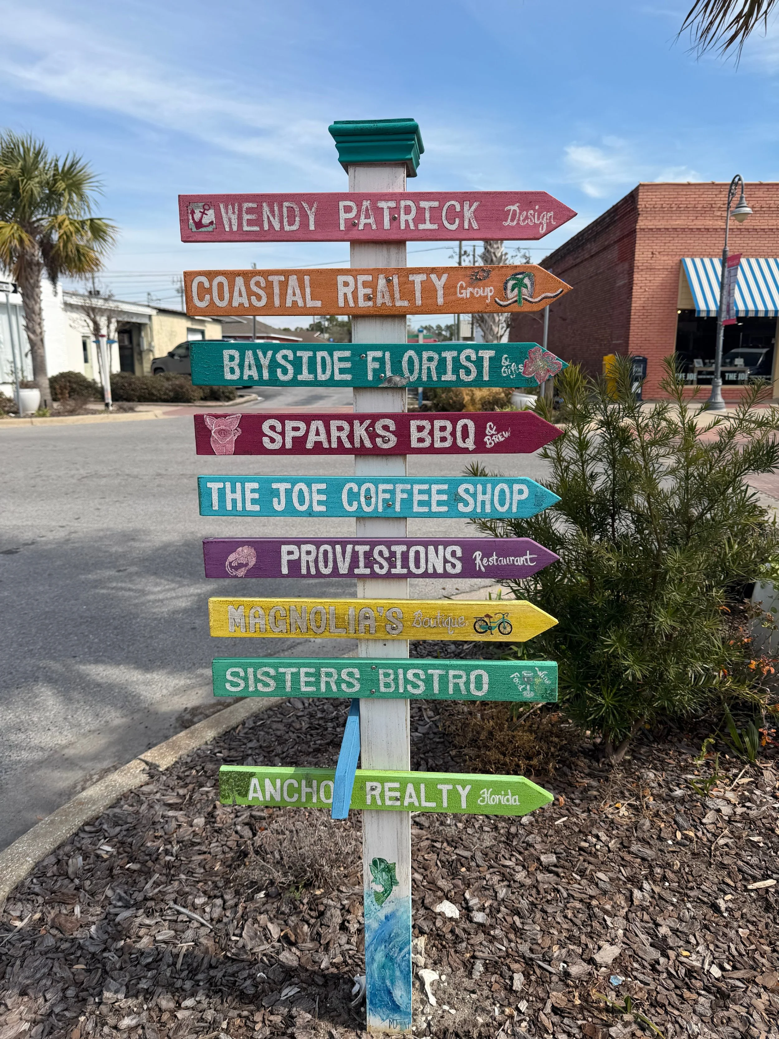 Colorful wooden directional signpost on sidewalk with multiple signs pointing in different directions, each with business or location names like Wendy Patrick Design, Coastal Realty, Bayside Florist, Spark's BBQ, The Joe Coffee Shop, Provisions Restaurant, Magnolia's Boutique, Sisters Bistro, and Anchor Realty Florida.