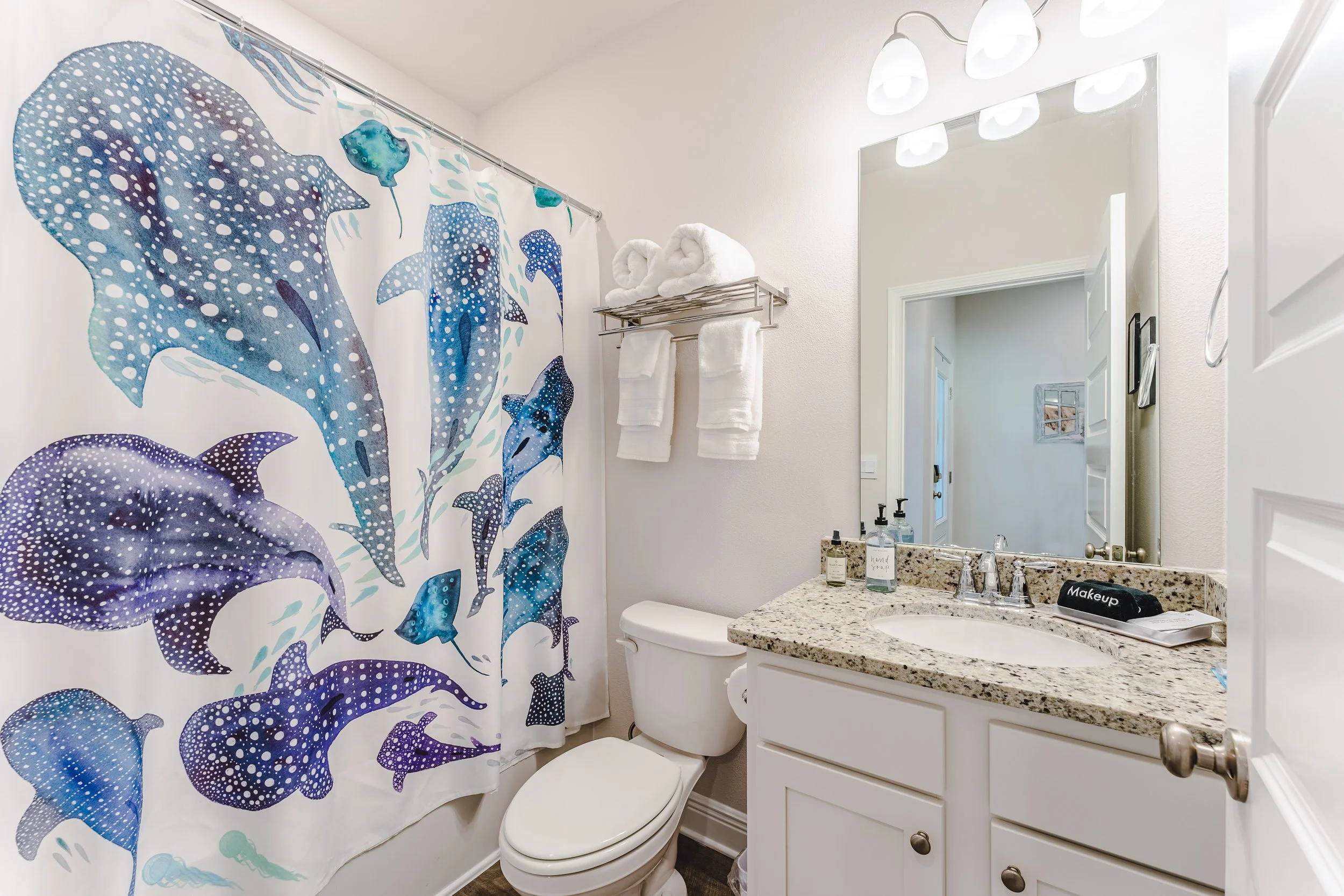 A small bathroom with a white vanity, granite countertop, and a large mirror. There are three wall-mounted lights above the mirror. On the wall, a towel rack holding two rolled towels and two folded towels. A shower curtain with a blue and purple dol