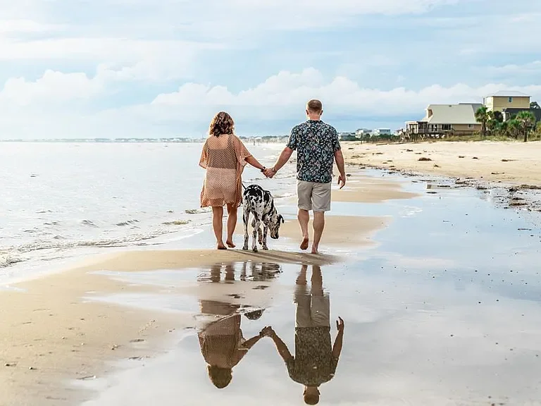 A couple walking hand-in-hand on the beach with their Dalmatian dog, near the shoreline with houses in the distance and a partly cloudy sky.