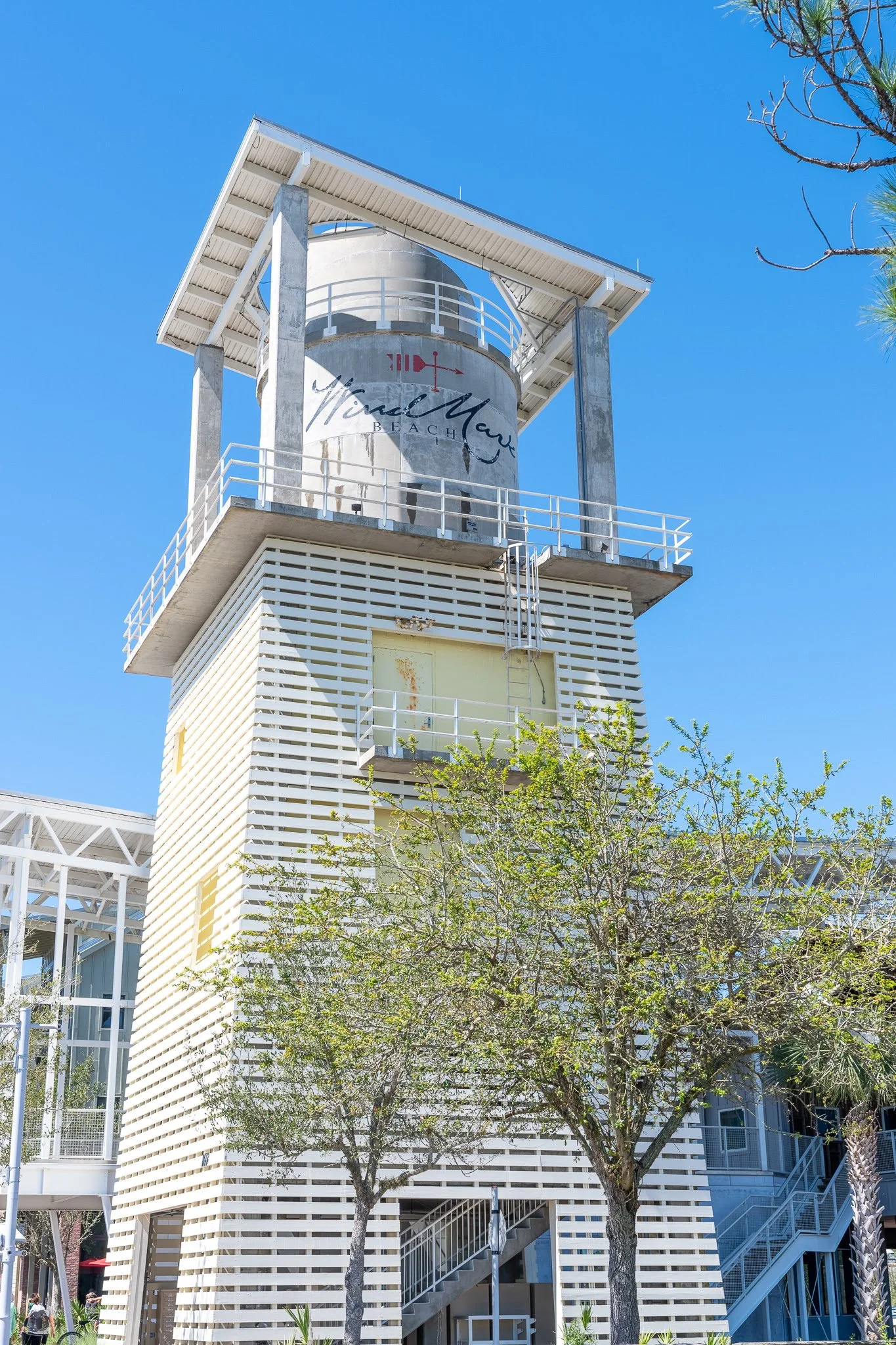 A tall structure with a lighthouse-like top, featuring the text 'Main Mouse Beach' on it, against a bright blue sky. Some trees and part of a building are visible at the base.