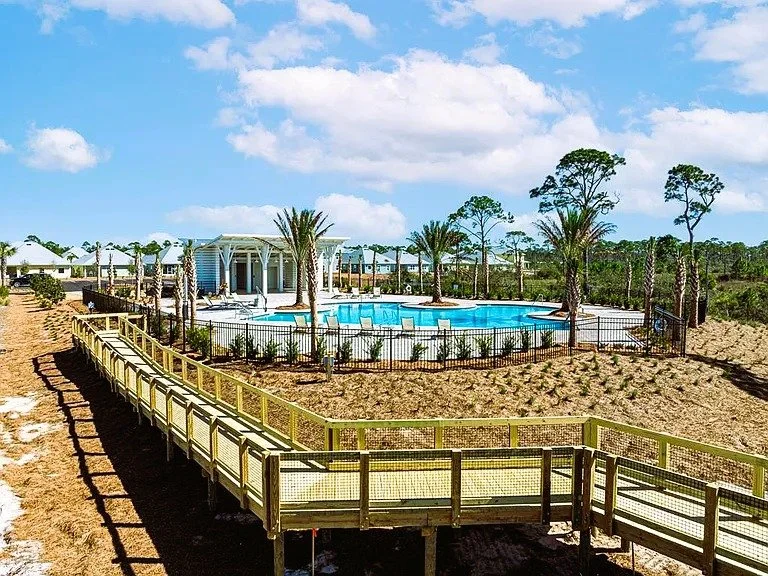 Resort-style swimming pool with surrounding palm trees, a pavilion, and a wooden walkway on sandy terrain under a partly cloudy sky.