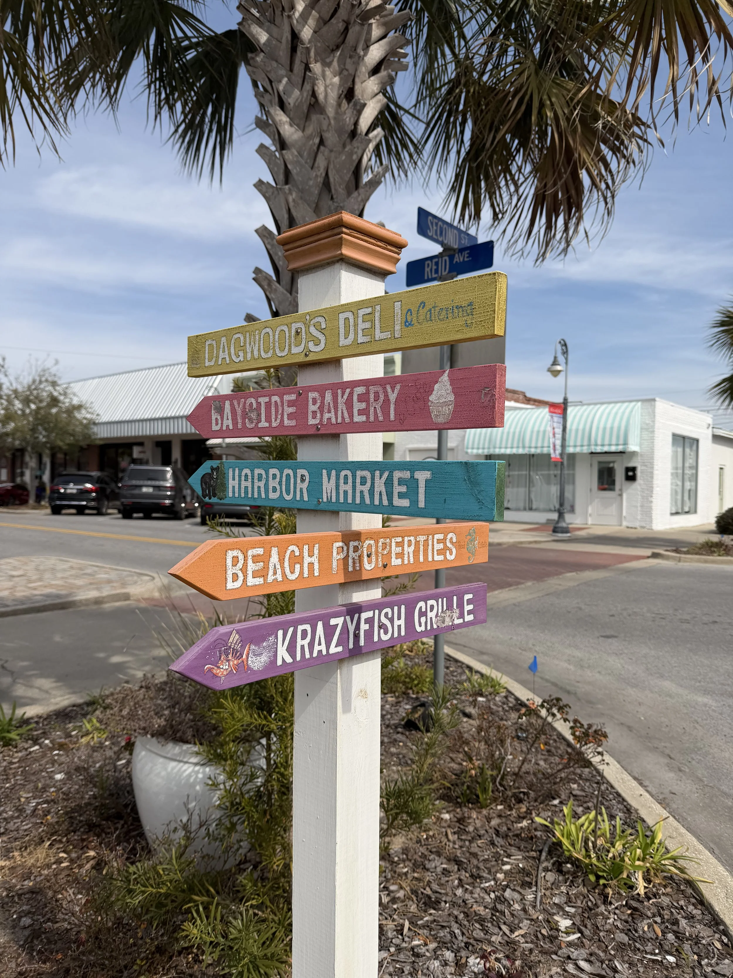 Colorful directional signpost in a small town with palm trees, showing locations like Dagwood's Deli, Bayside Bakery, Harbor Market, Beach Properties, and Krazyfish Grille.