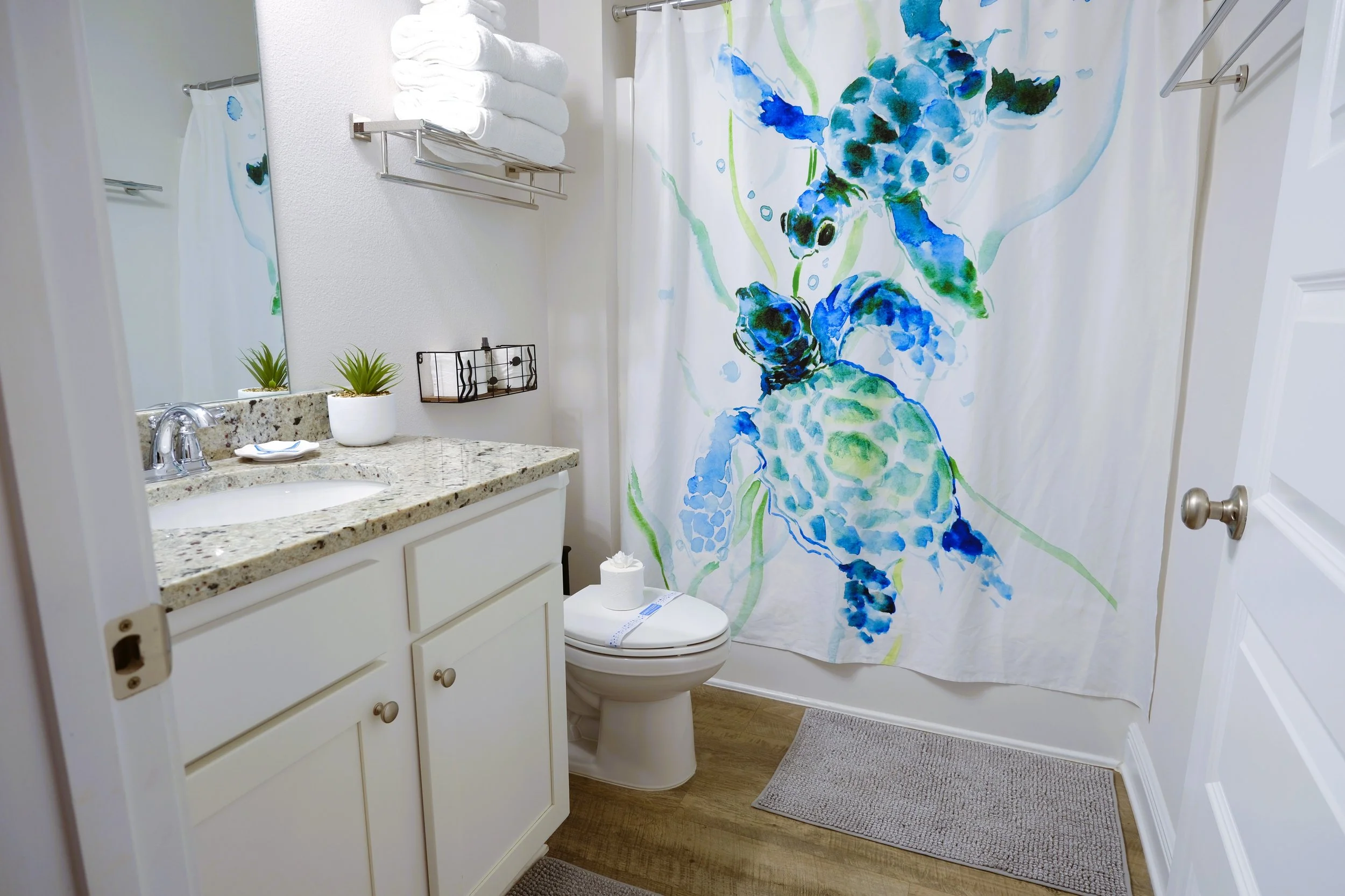 A small bathroom with a white vanity, granite countertop, and a large mirror. There are three wall-mounted lights above the mirror. On the wall, a towel rack holding two rolled towels and two folded towels. A shower curtain with a blue and purple dol
