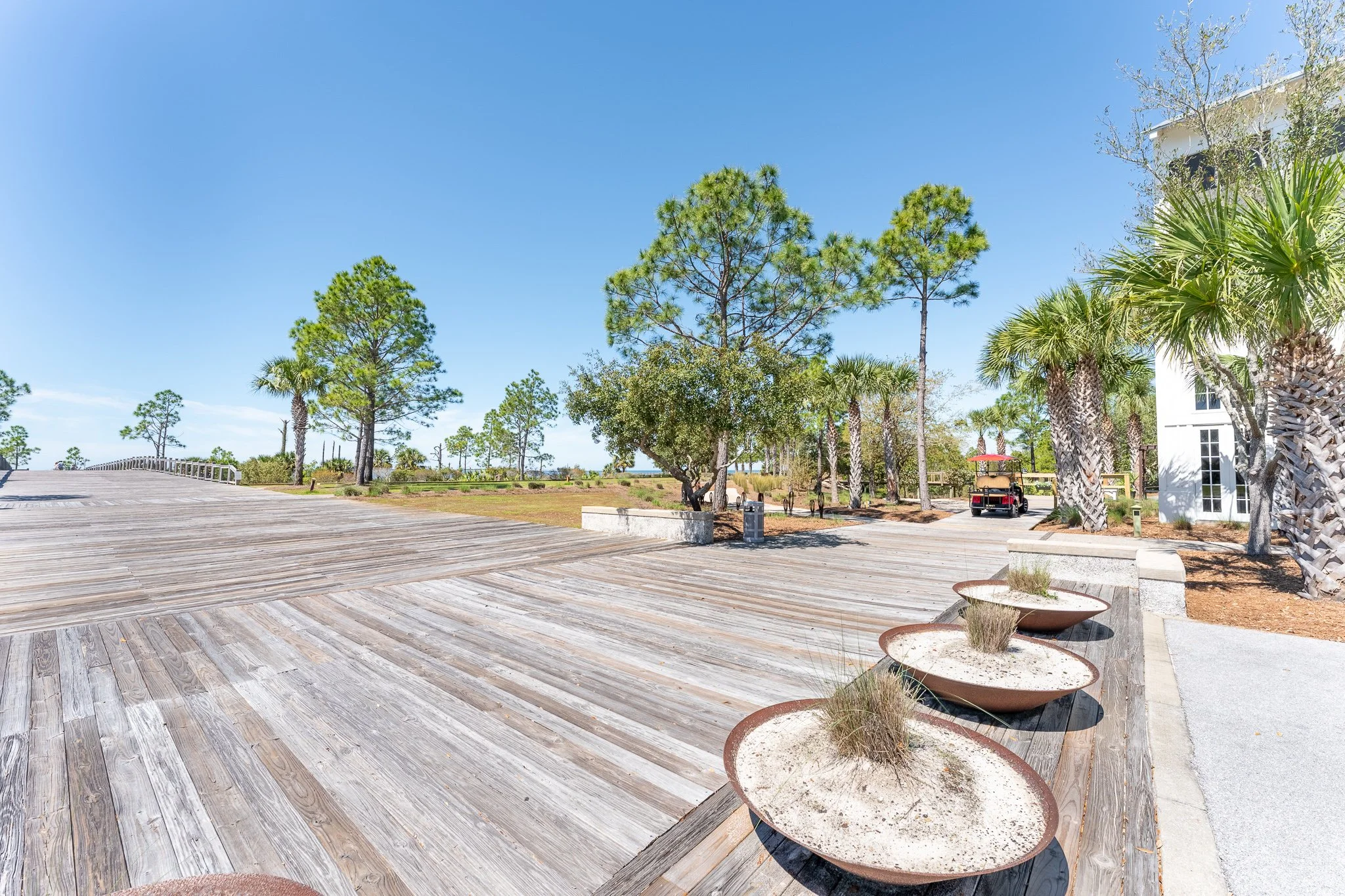 Sunlit outdoor wooden deck with planters, surrounded by palm and pine trees, and a golf cart parked near a white building.