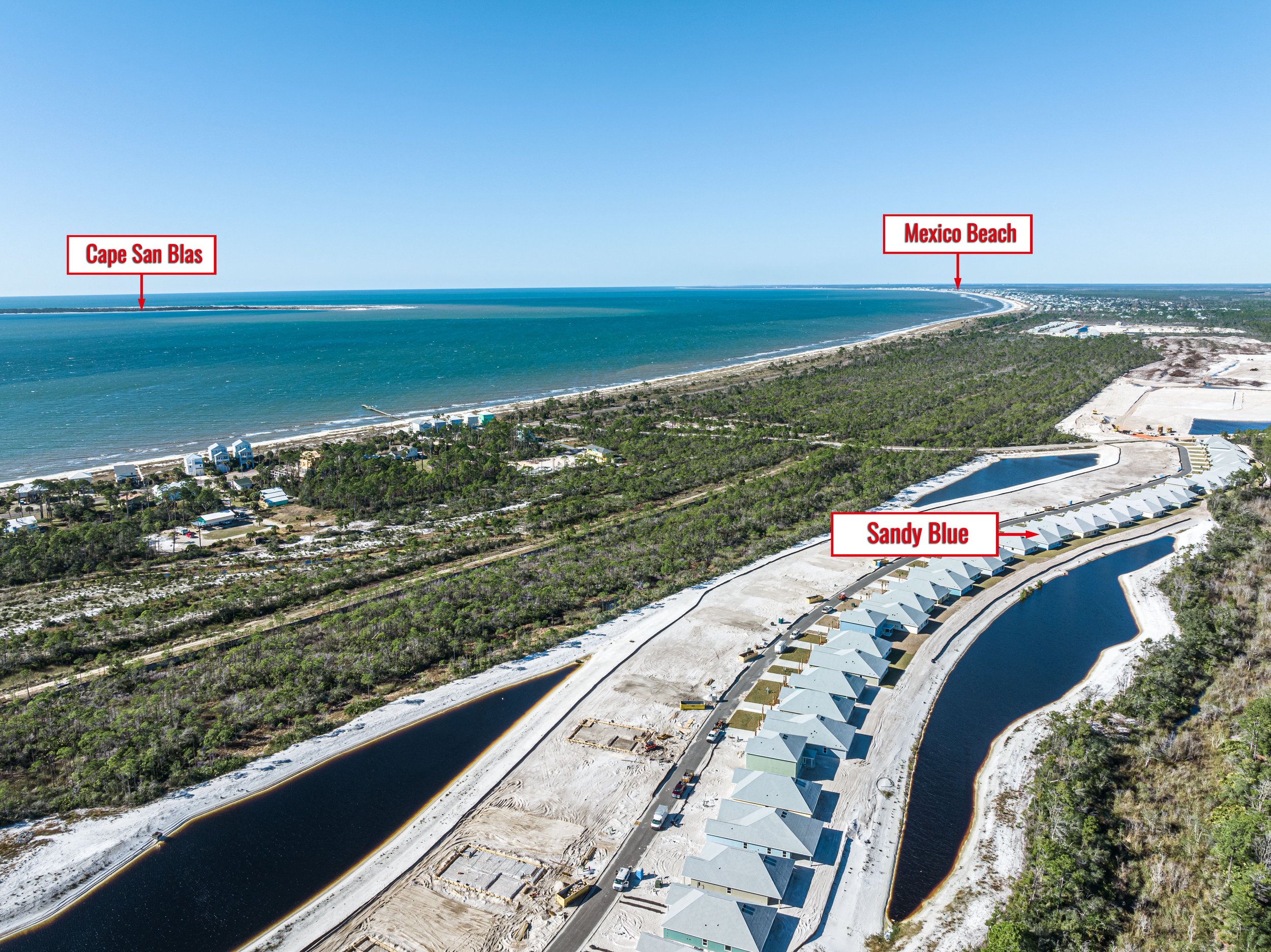 An aerial view of a coastal area showing the beach and ocean with labels pointing to Cape San Blas and Mexico Beach in the distance. There are also houses built along a canal, with one section labeled Sandy Blue. The landscape includes greenery, construction areas, and water channels.