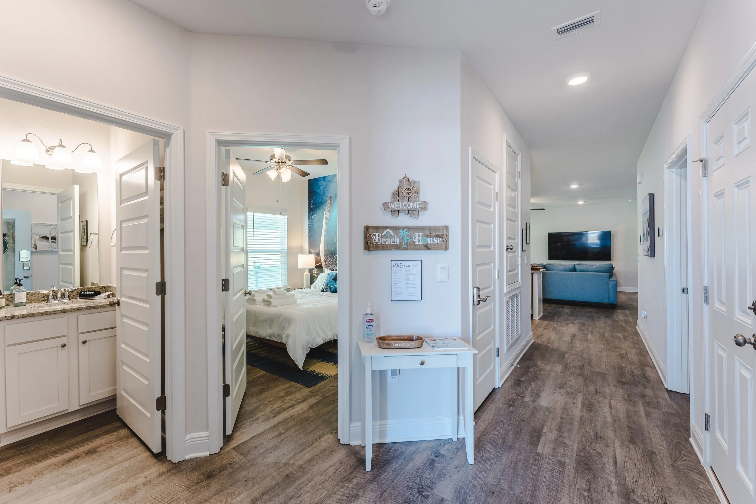 Interior of a vacation rental with a view into a bedroom, bathroom, and living room, featuring white walls, wood flooring, and beach-themed decorations.