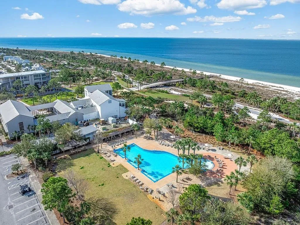 Aerial view of a resort with a large swimming pool, surrounded by palm trees and lounge chairs, near a white building, with the beach and ocean in the background.