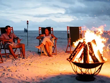 Two people sitting on beach chairs near a campfire on the beach, with the ocean and cloudy sky in the background.