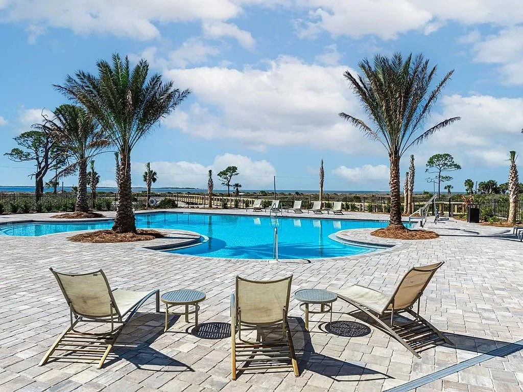 A swimming pool area with palm trees, lounge chairs, and a view of the ocean and sky with clouds.