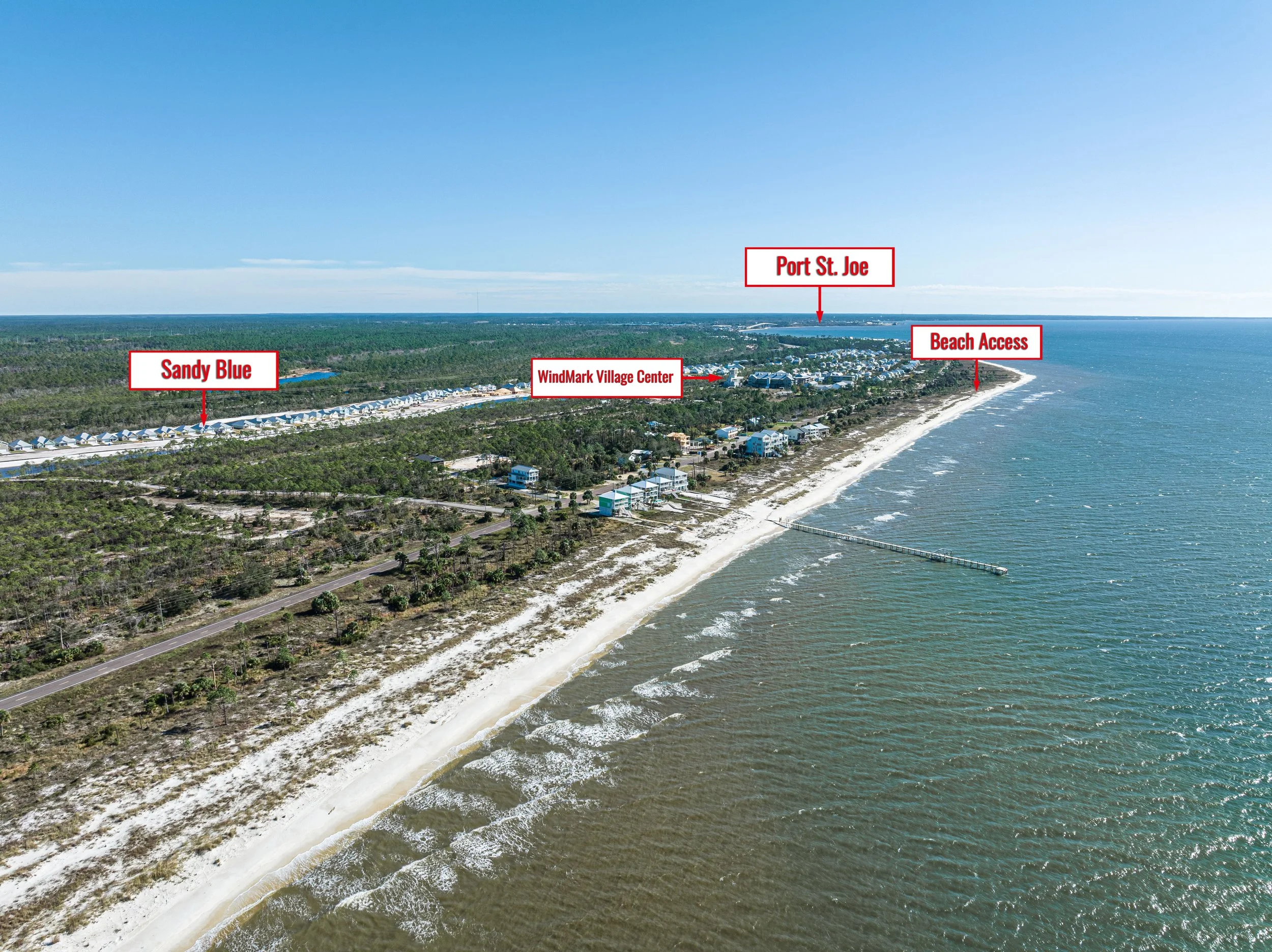 An aerial view of a coastal area with beach access, labeled landmarks including Sandy Blue, WindMark Village Center, Port St. Joe, and the beach. The beach has a long pier extending into the water, and the landscape features sandy shores, ocean waves, and green vegetation inland.