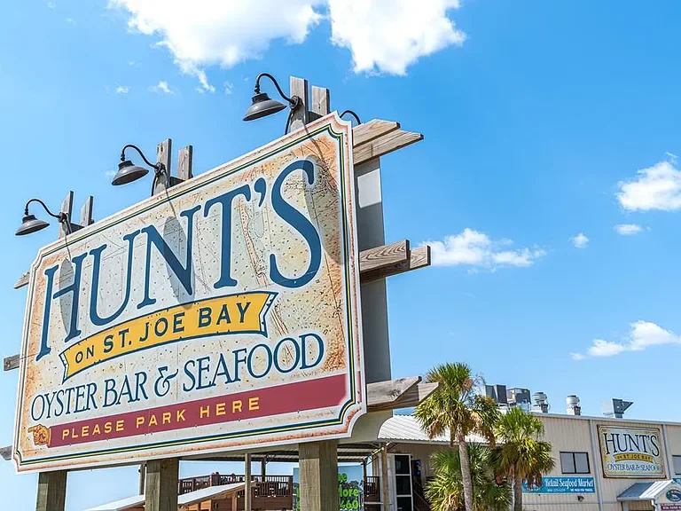 Sign for Hunt's Oyster Bar & Seafood on St. Joe Bay with a clear blue sky, palm trees, and buildings in the background.