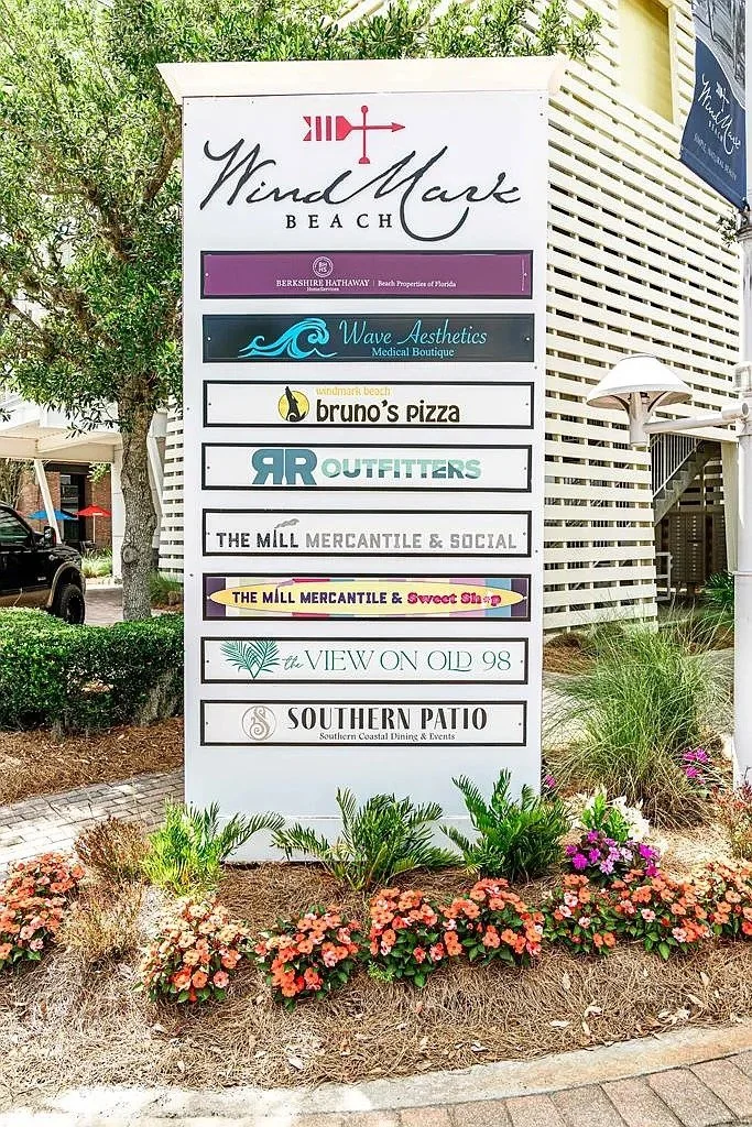 A vertical signboard listing multiple businesses at WindMark Beach, including Berkshire Hathaway, Wave Aesthetics, Bruno's Pizza, RR Outfitters, The Mill Mercantile & Social, The Mill Mercantile & Sweet Shop, The View on Old 98, and Southern Patio, with flowers and greenery at the base.