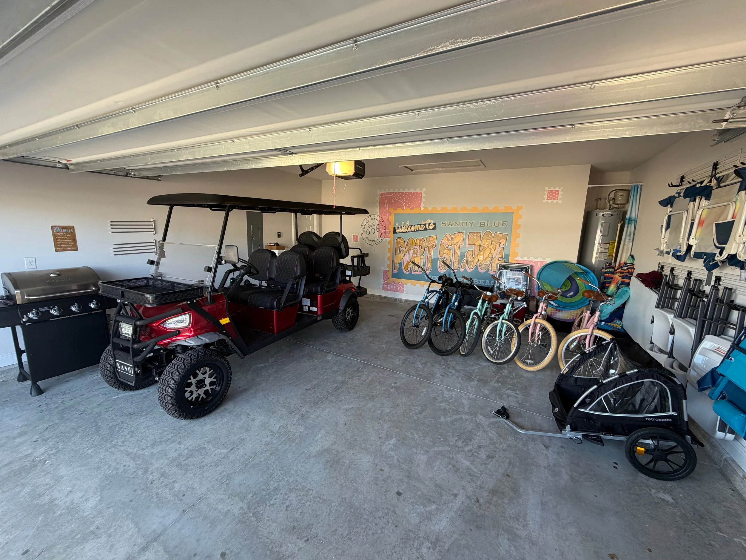 Garage with a red golf cart, several bicycles, a black bike trailer, a propane grill, and beach accessories such as floaties and chairs, with a colorful wall mural that says "Welcome to Sandy Blue Pop Stage."
