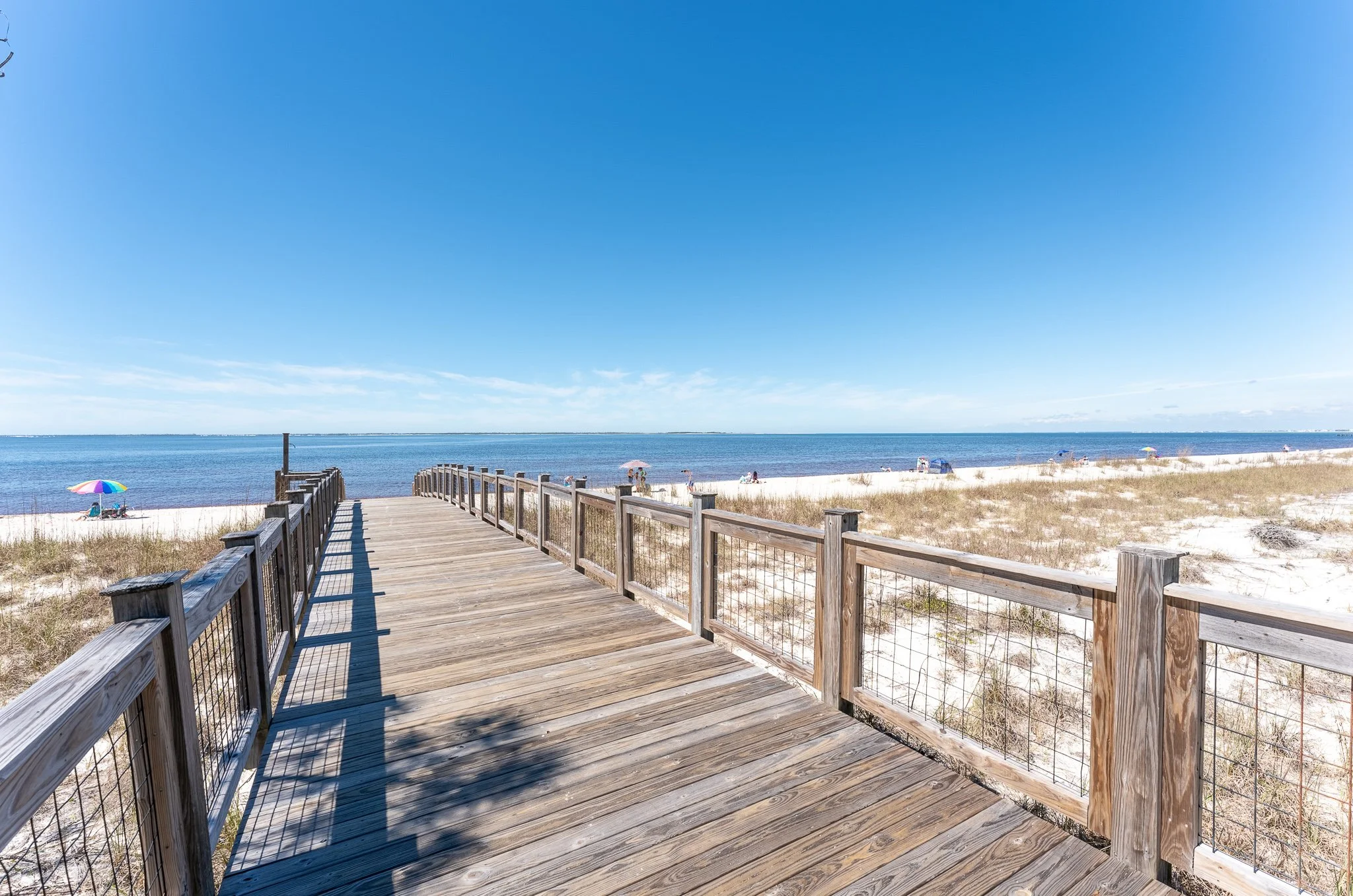 Wooden boardwalk leading to a sandy beach with people under umbrellas, blue sky and ocean in the background.