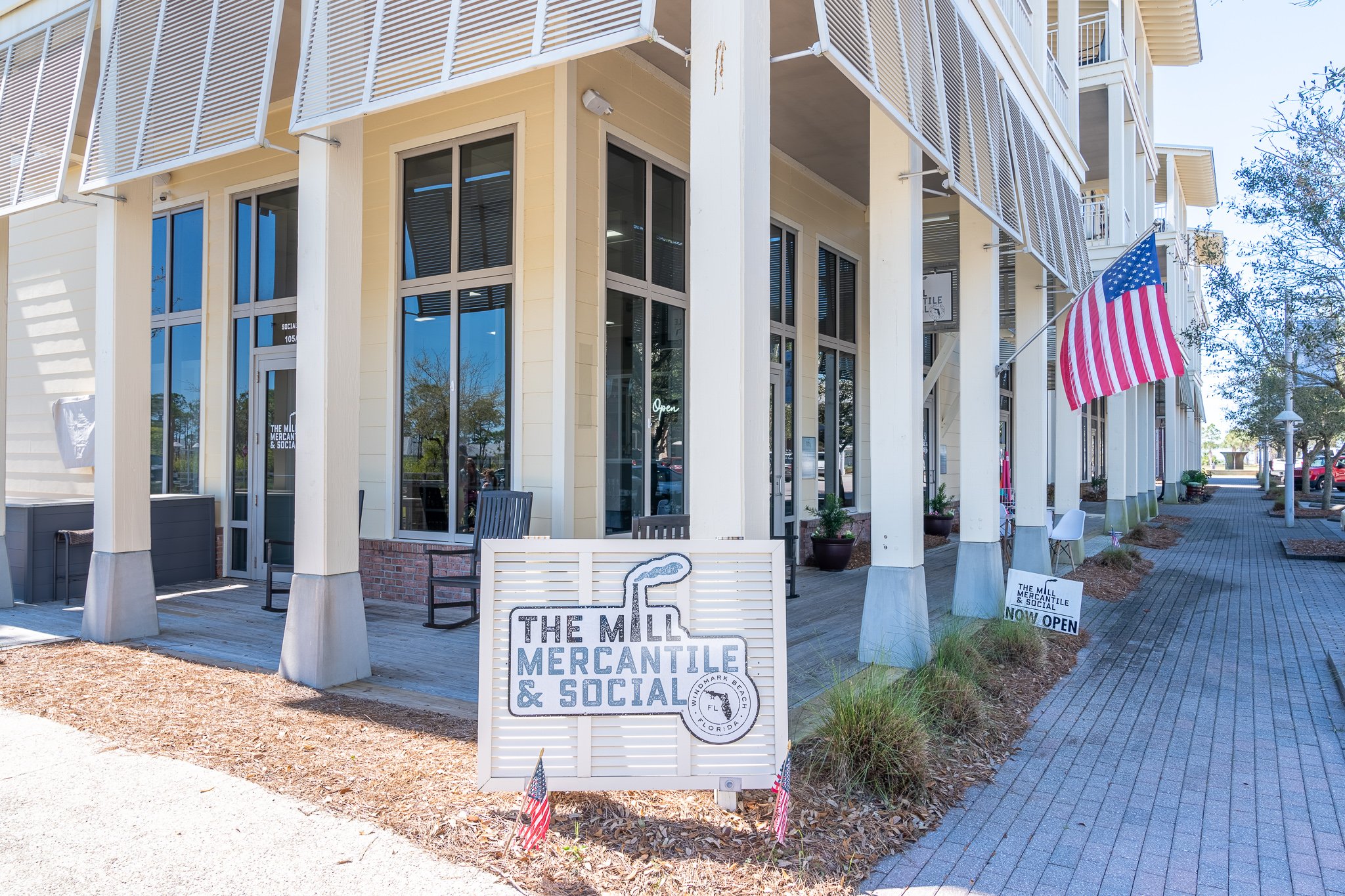 Exterior view of a building with large windows, a sign that reads 'The Mill Mercantile & Social', and an American flag outside. There are chairs and potted plants along the sidewalk, with a small sign indicating the shop is now open.