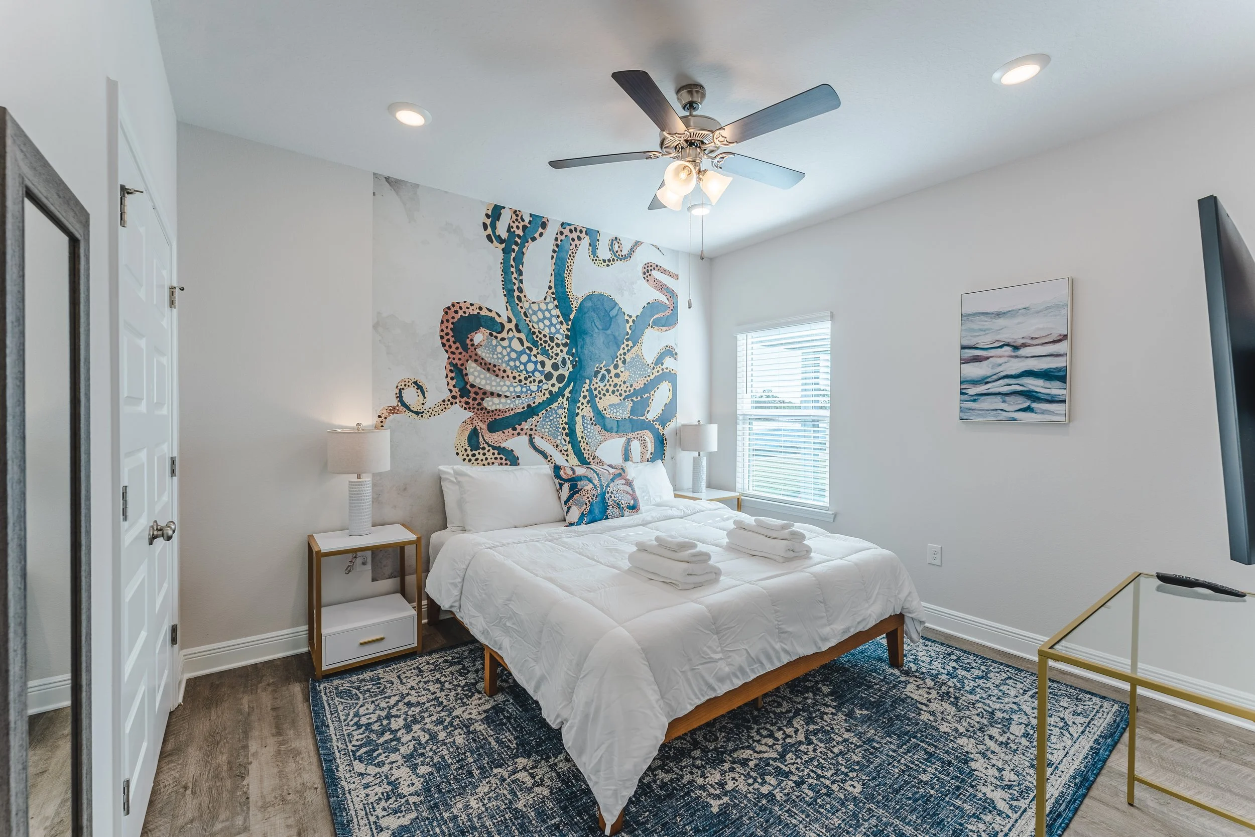 A modern bedroom with white walls, a bed with white linens, two bedside tables with lamps, a ceiling fan, a window with blinds, a wall painting of ocean waves, and a blue patterned rug.