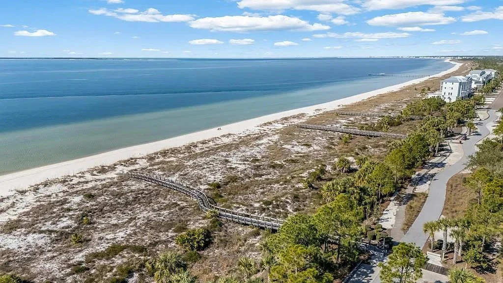 A coastal beach with white sand, a boardwalk, and a beachside neighborhood with houses and palm trees.