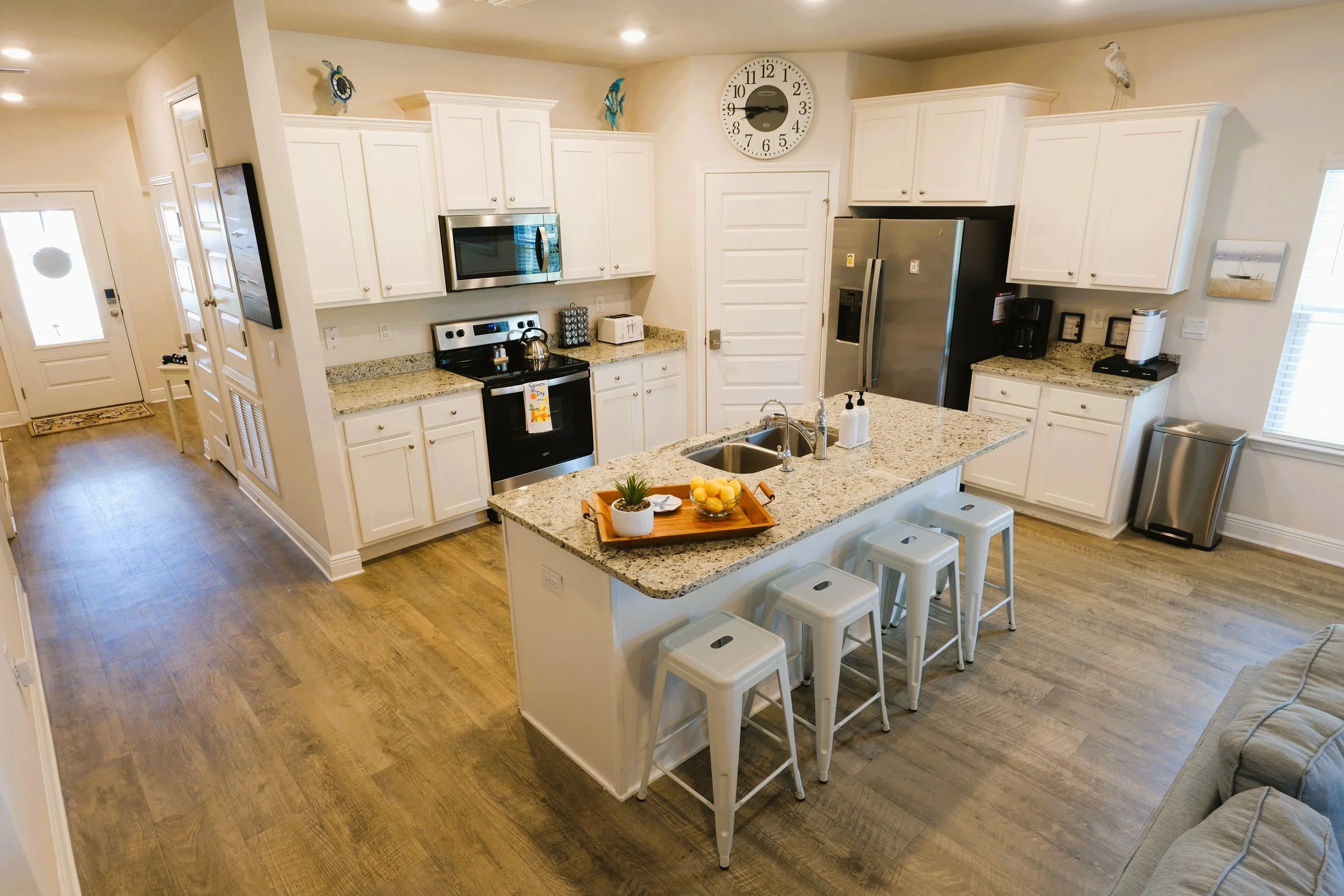 Bright kitchen with white cabinets, stainless steel appliances, granite countertops, and a kitchen island with four white barstools. There are recessed ceiling lights, a window with blinds, and a wall clock.