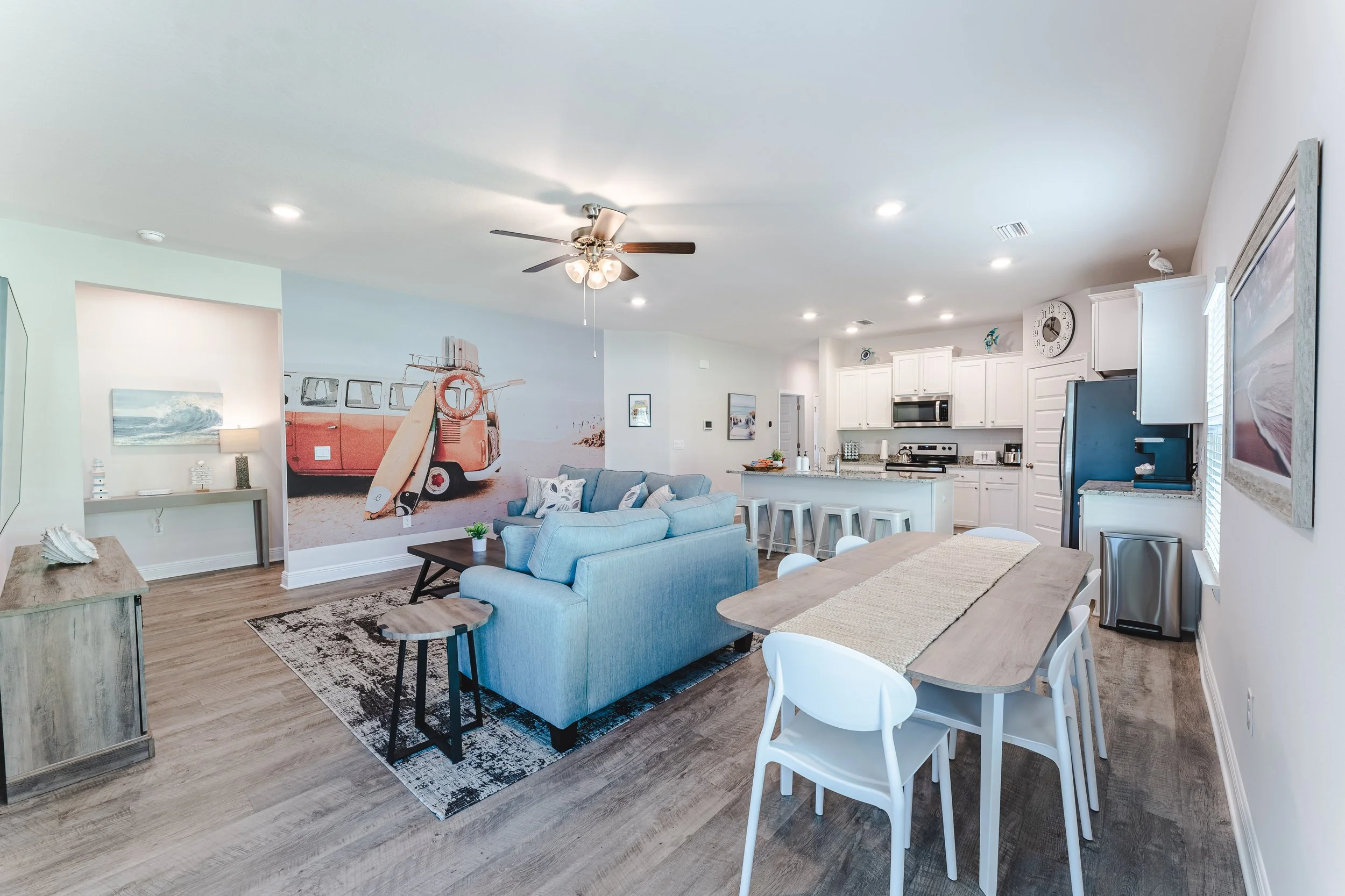 Open-concept living room and kitchen with ocean wave and surfboard mural, light hardwood flooring, gray sofas, wooden dining table with white chairs, white kitchen cabinets, and stainless steel appliances.