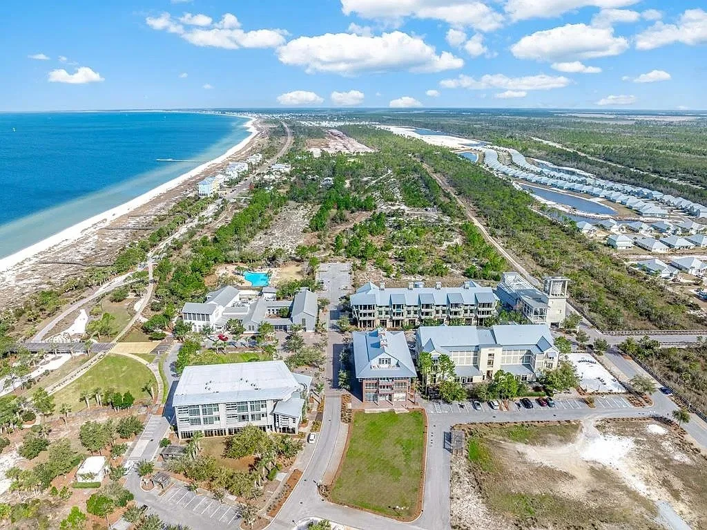 Aerial view of a coastal area with a beach, buildings, parking lots, and water channels, with green trees and fields.