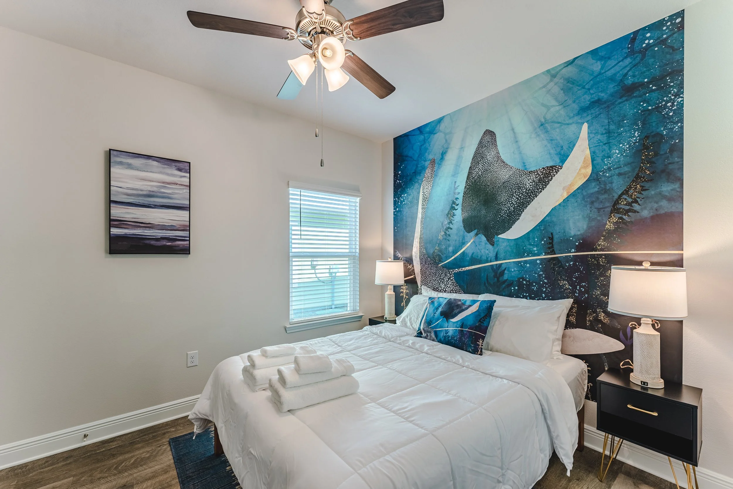 A bedroom with a white bed, ocean-themed mural, and ceiling fan with lights.