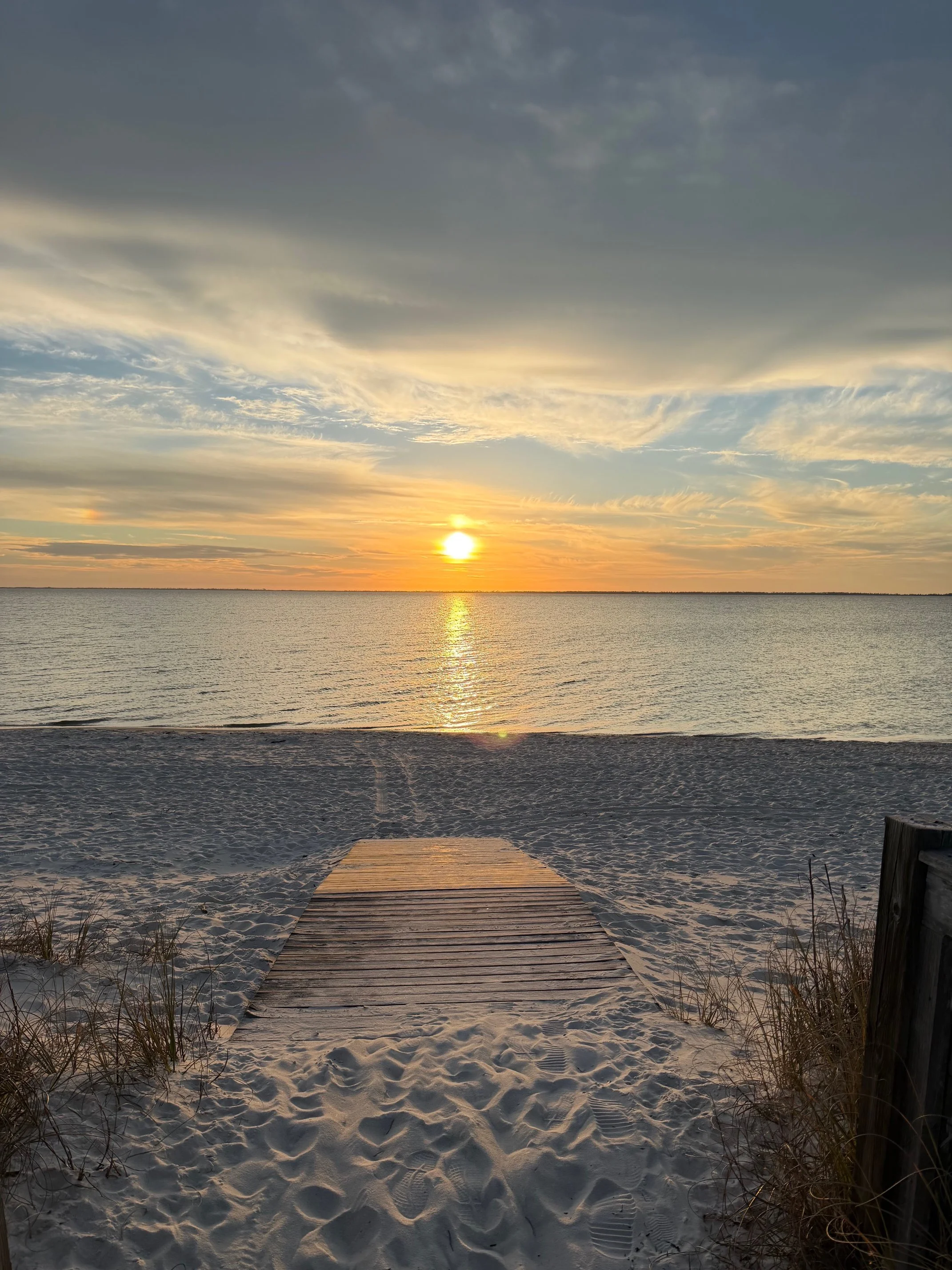 Sunset over the ocean viewed from a sandy beach with a wooden pathway leading to the water, clouds in the sky, and a calm sea.