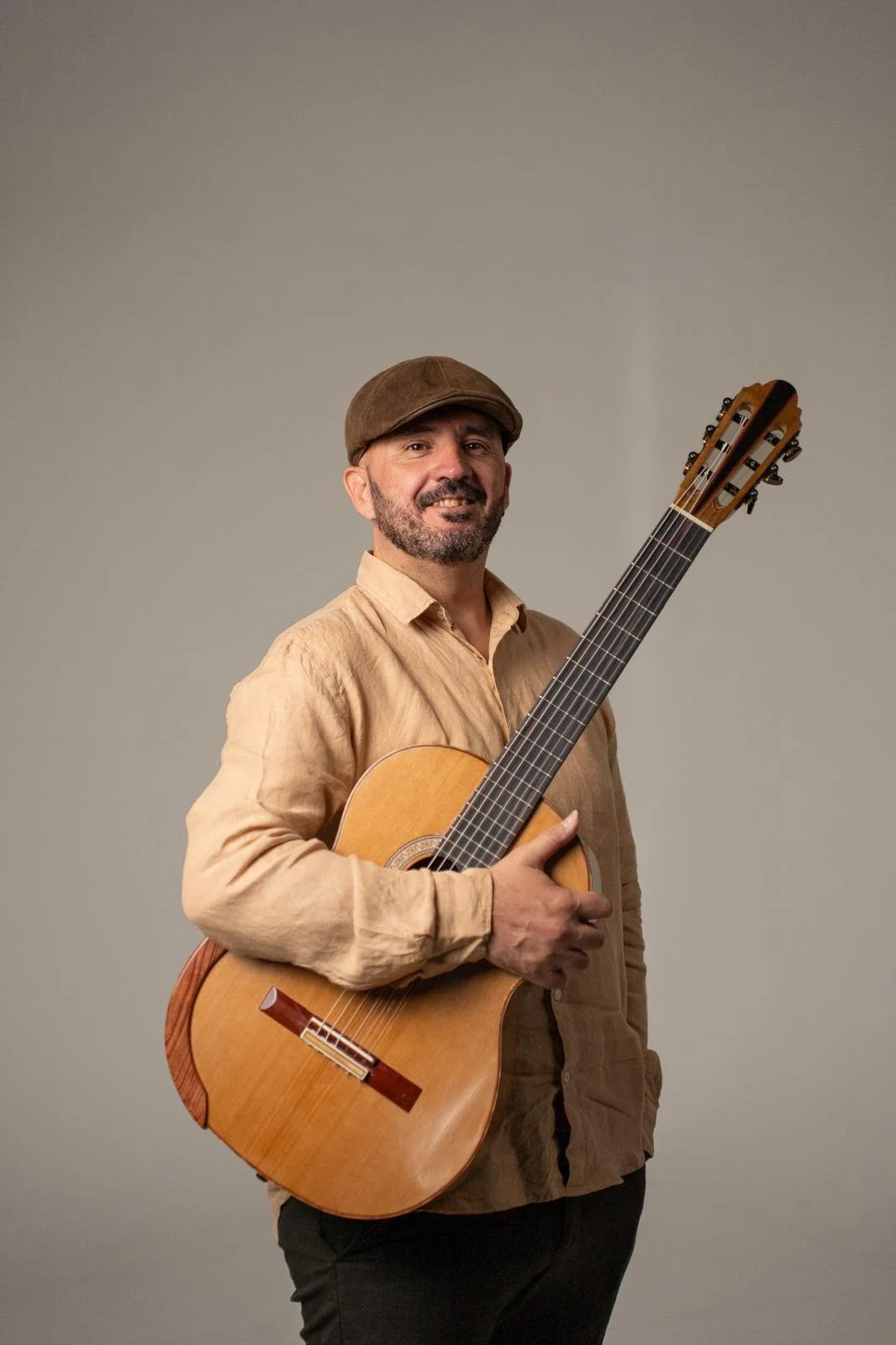 Hombre con barba, sombrero y camisa beige sosteniendo una guitarra clásica, sonriendo, en un fondo gris.
